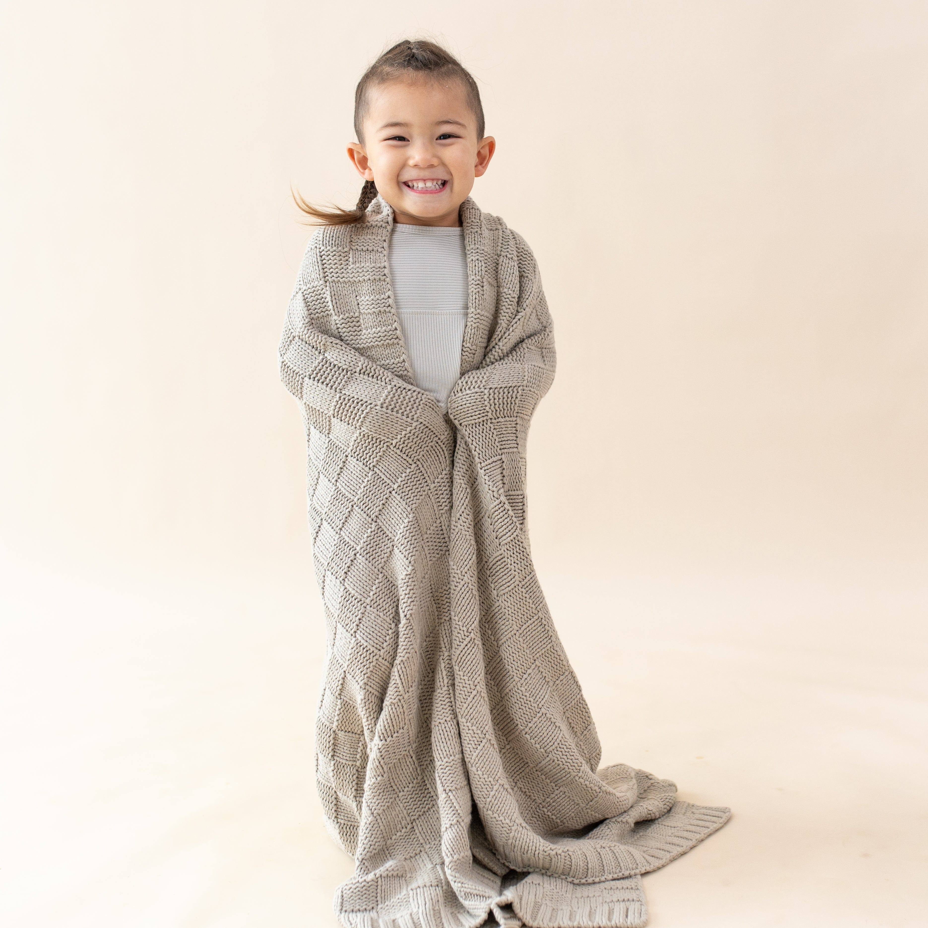 Girl wrapped in Kyte Baby Chunky Knit Basket Weave Toddler Blanket in Almond