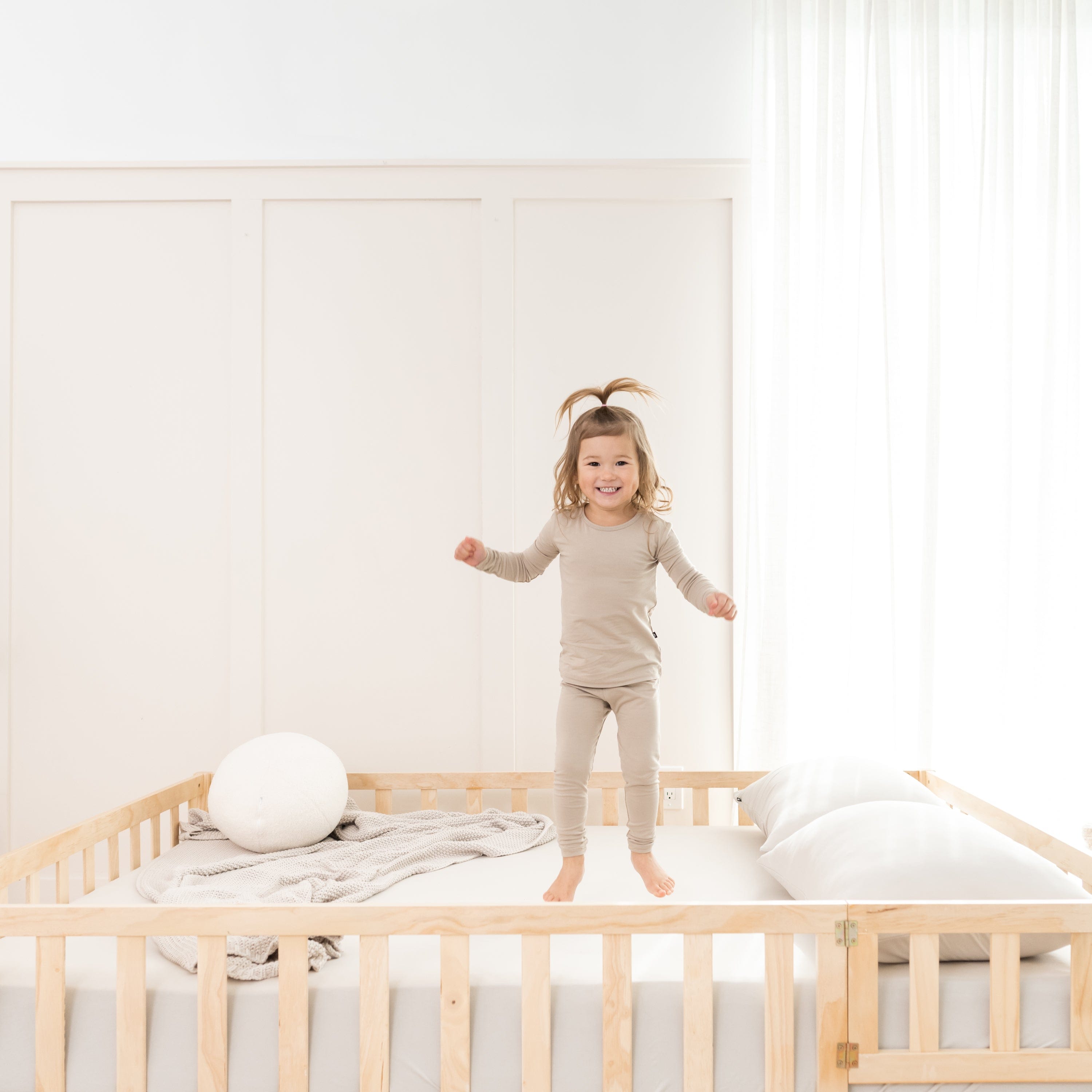 Toddler jumping on bed made with fitted sheet wearing two piece Kyte Baby Long Sleeve Pajamas in Almond