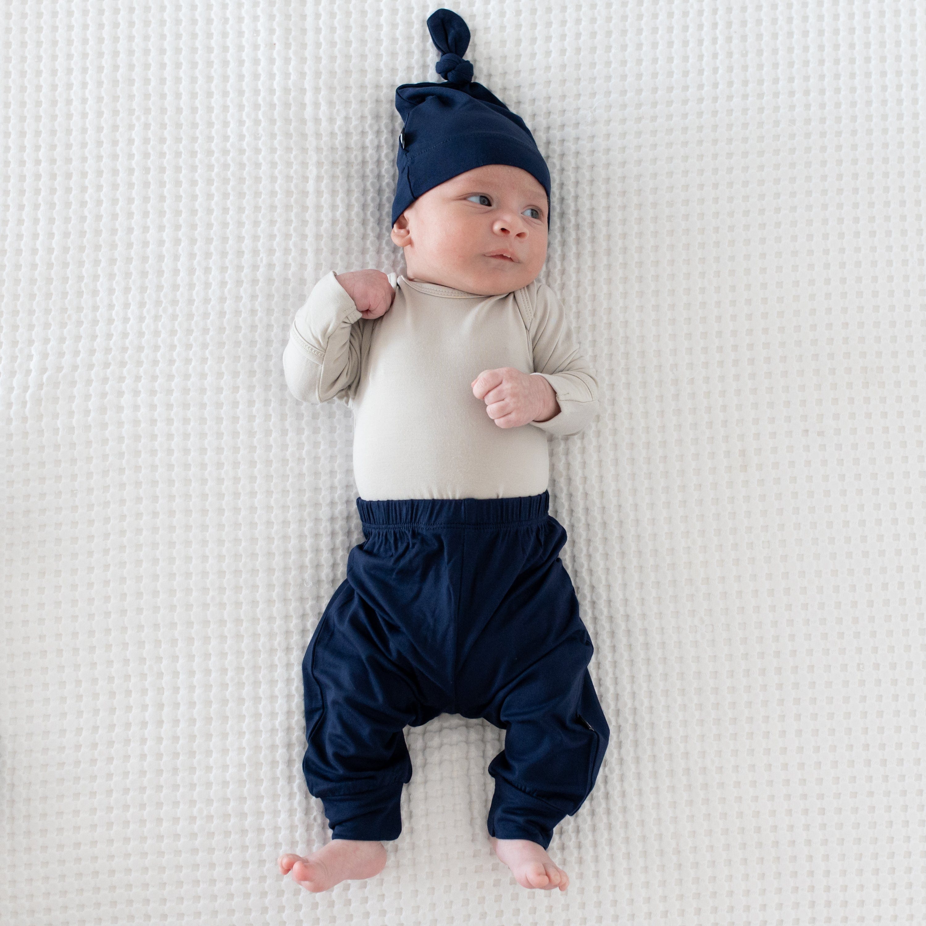 Newborn wearing Kyte Baby Knotted Cap in Navy with matching pants and a Long Sleeve Bodysuit in Oat