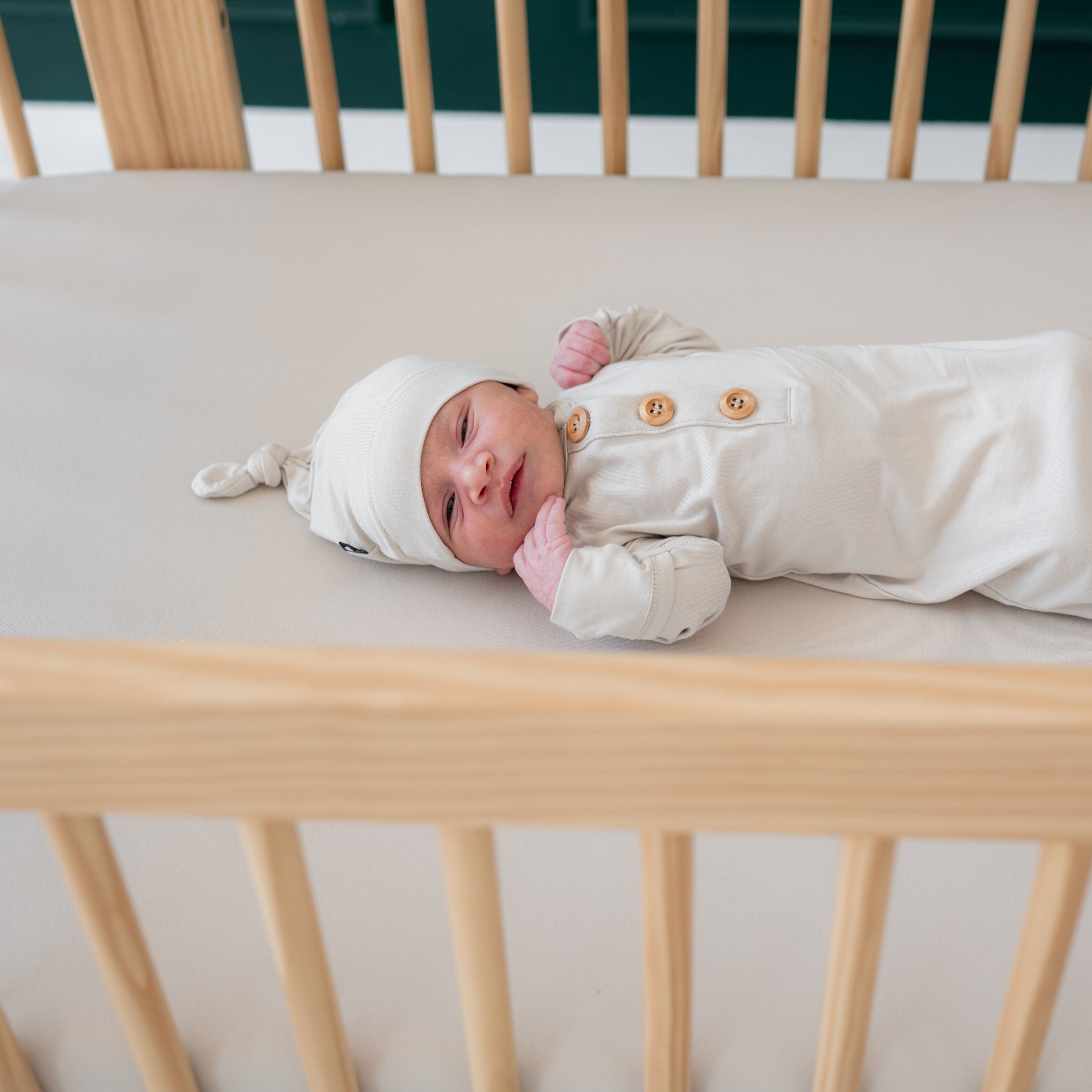 Baby laying in wooden crib and wearing Kyte Baby Knotted Gown with Hat Set in Oat