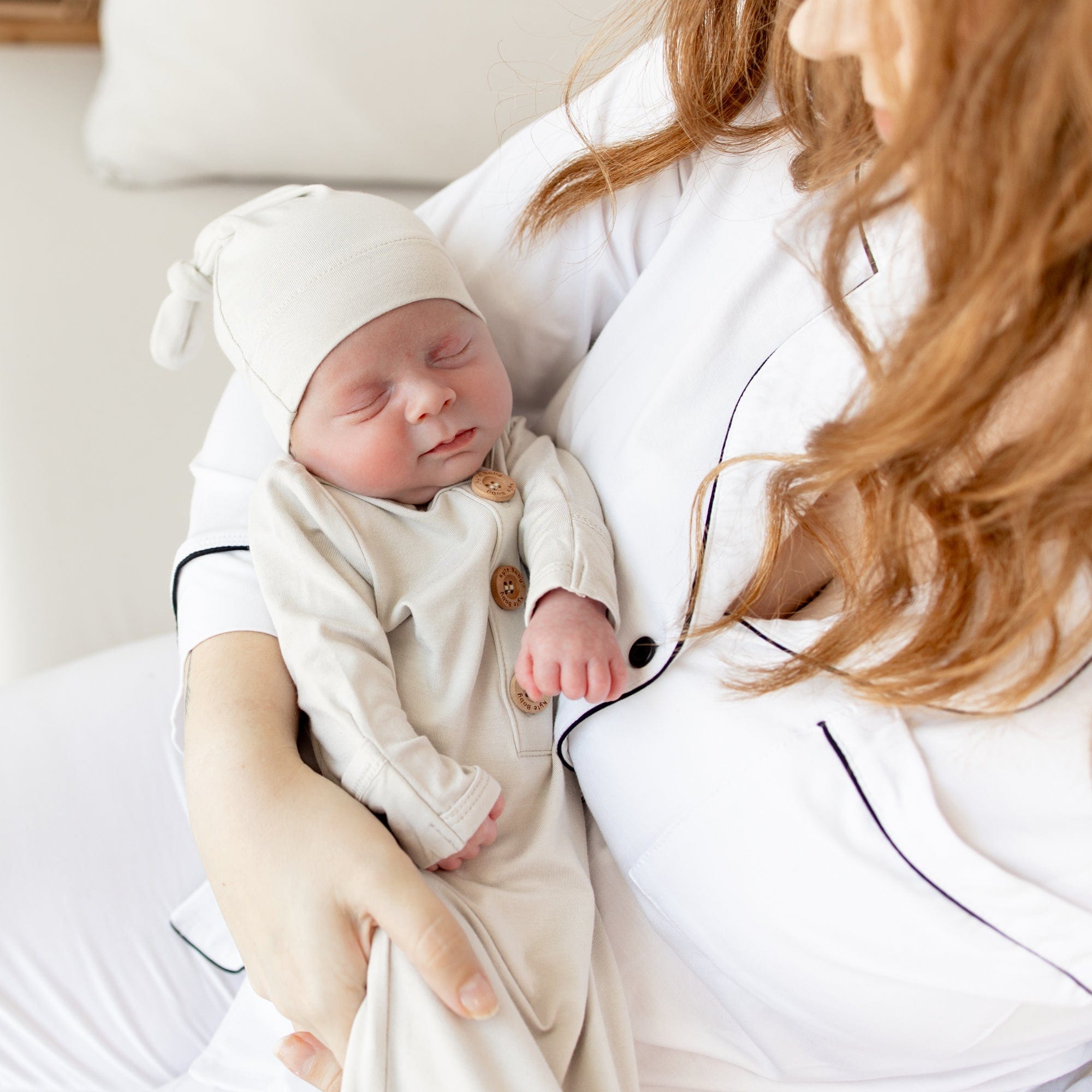 Newborn sleeping in mother's arms while wearing Kyte Baby Knotted Gown with Hat Set in Oat