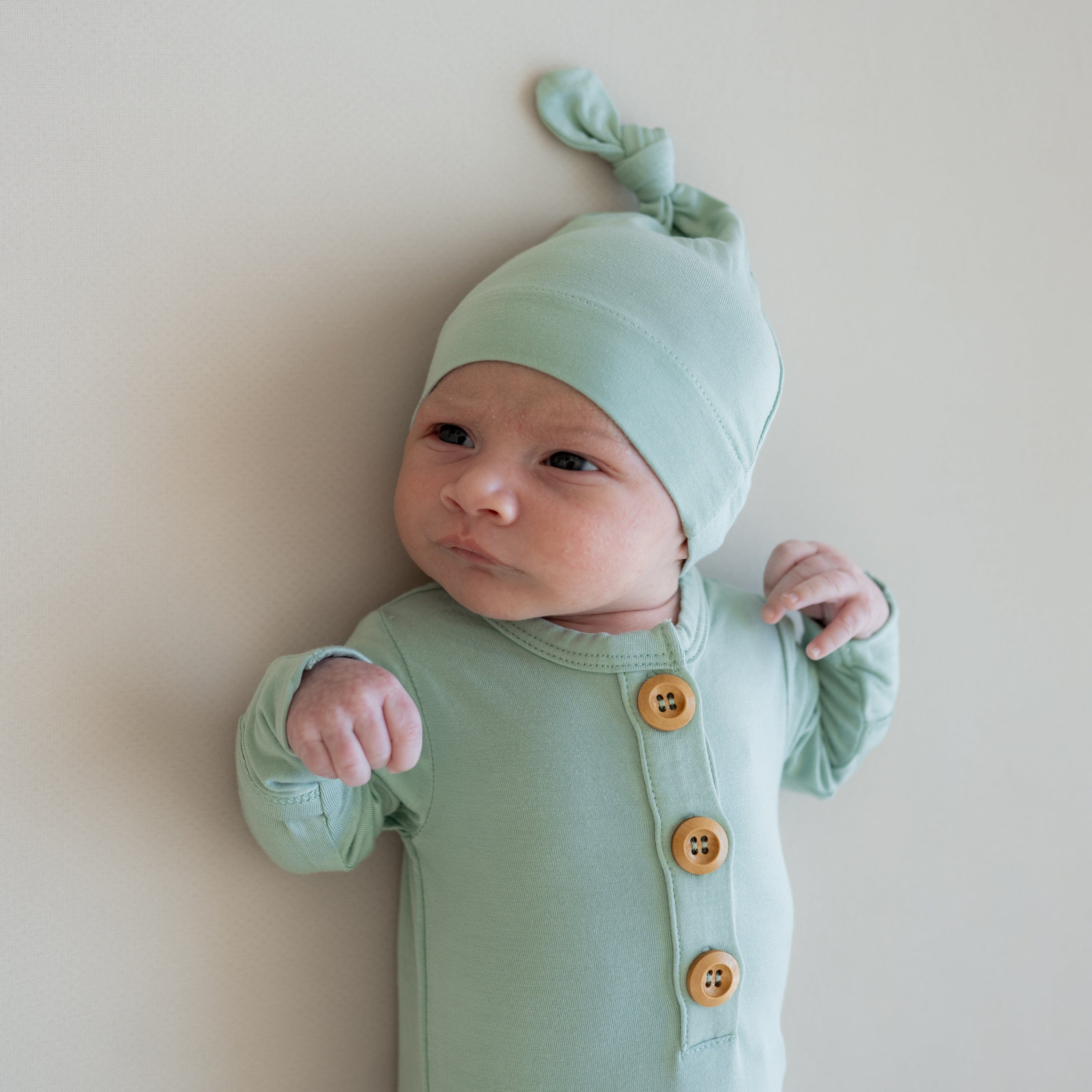 Close up of newborn wearing Kyte Baby Knotted Gown with Hat Set in Sage while laying on a crib sheet in Oat