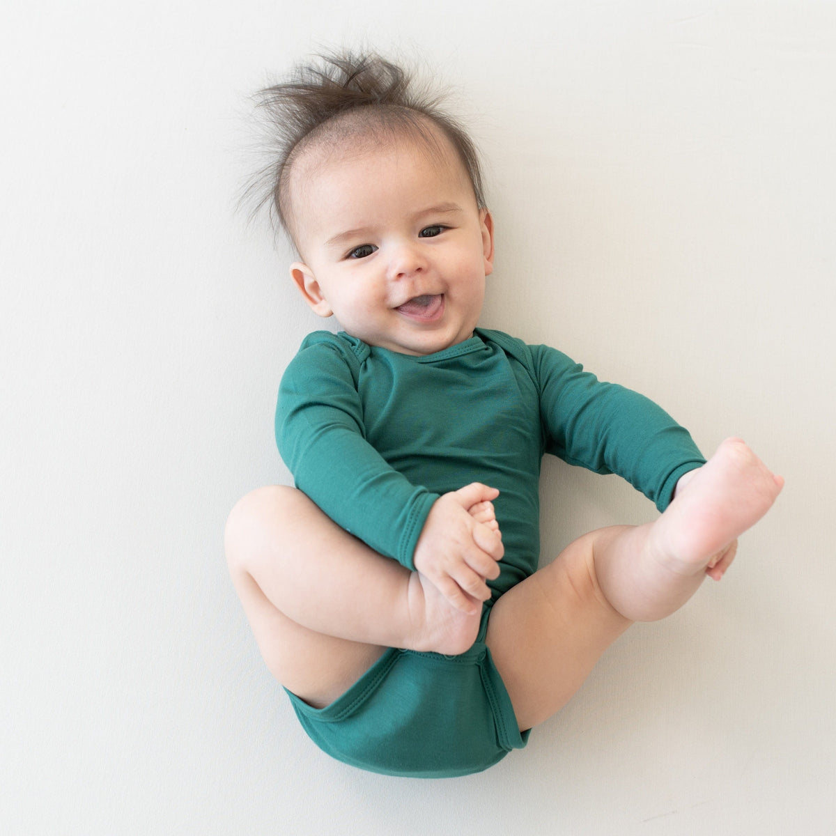 Smiling baby holding feet and wearing Kyte Baby Long Sleeve Bodysuit in Emerald