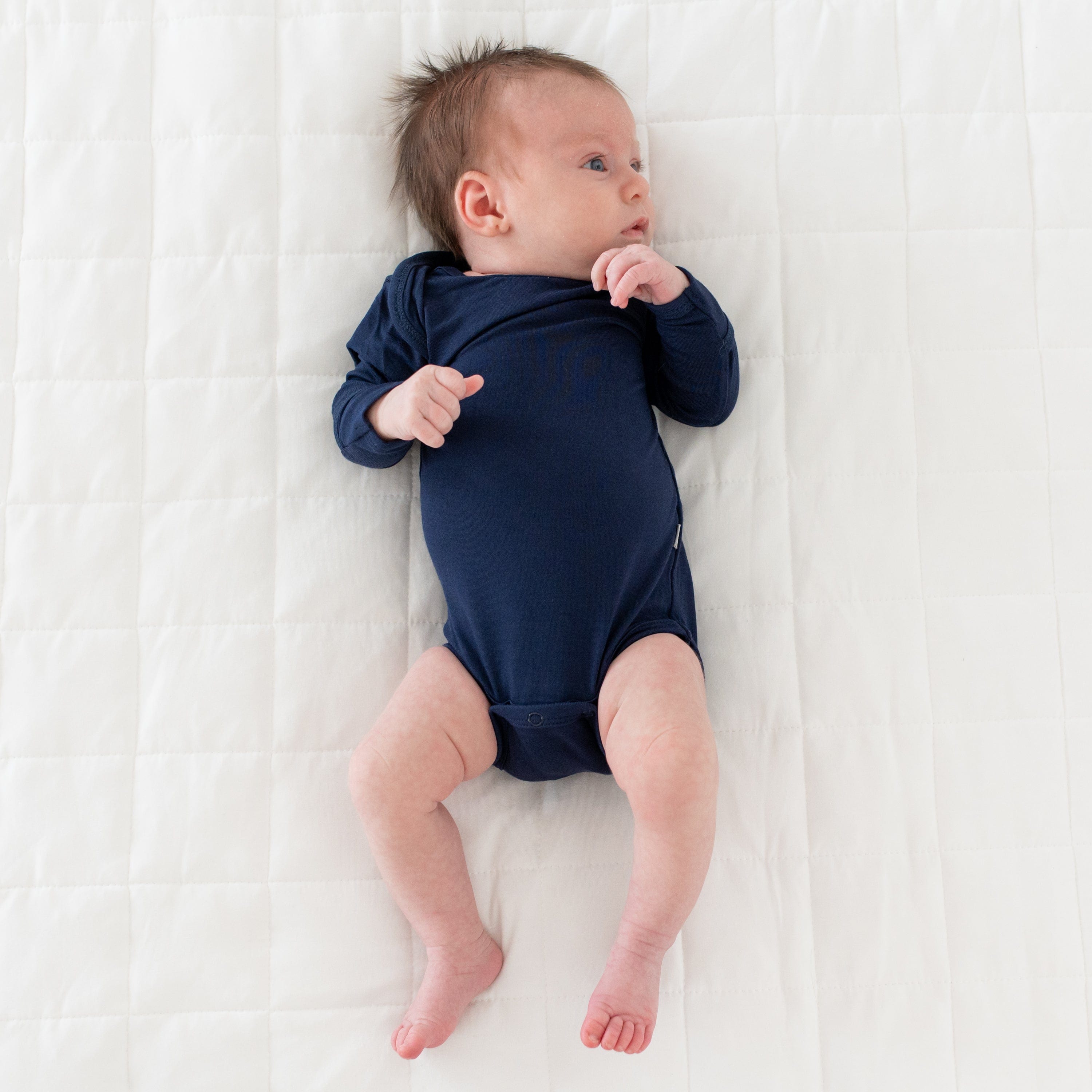 Newborn wearing Kyte Baby Long Sleeve infant Bodysuit in Navy while laying on its back on a the quilted side of a blanket in Oat