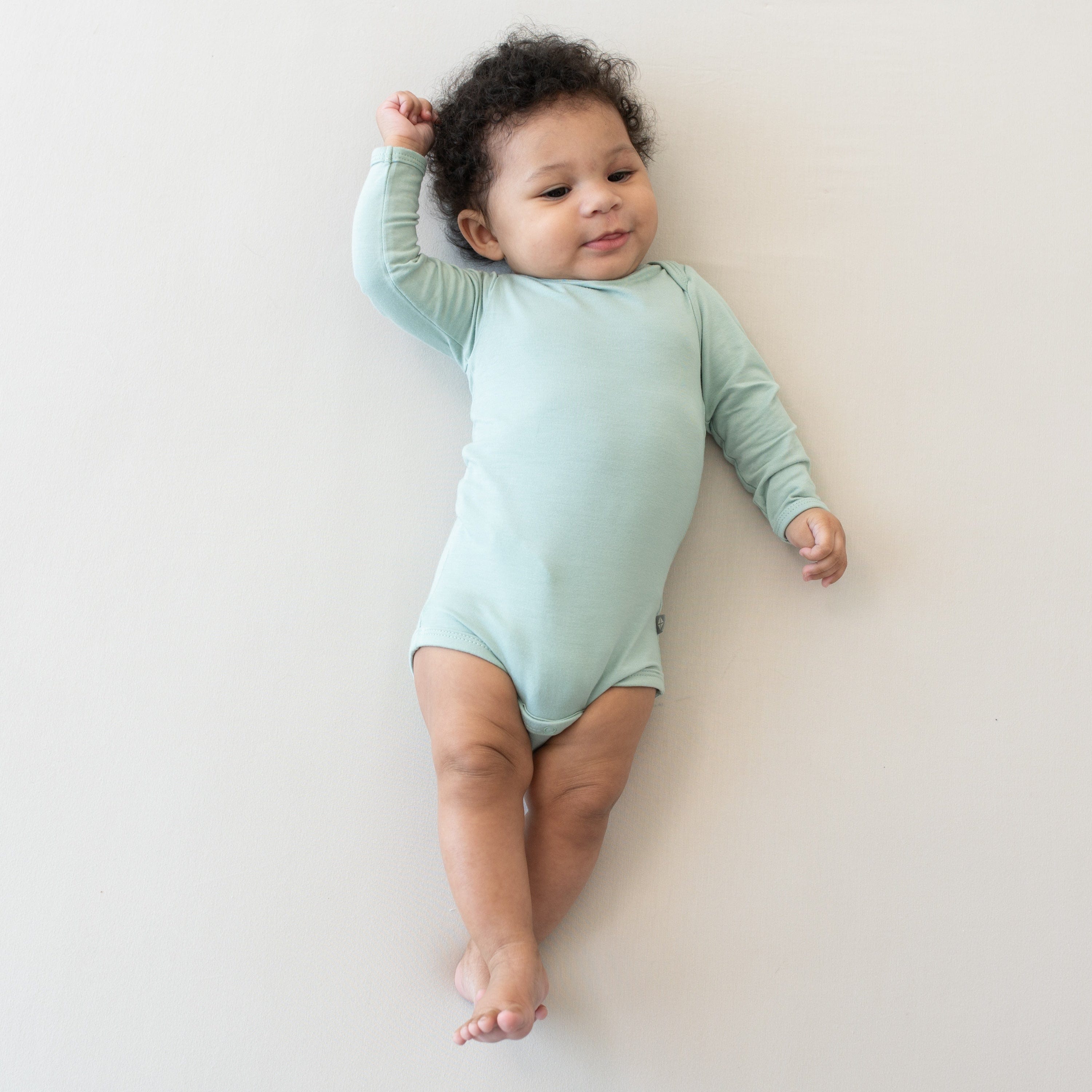 Infant wearing Kyte Baby Long Sleeve Bodysuit in Sage with legs crossed and one arm raised