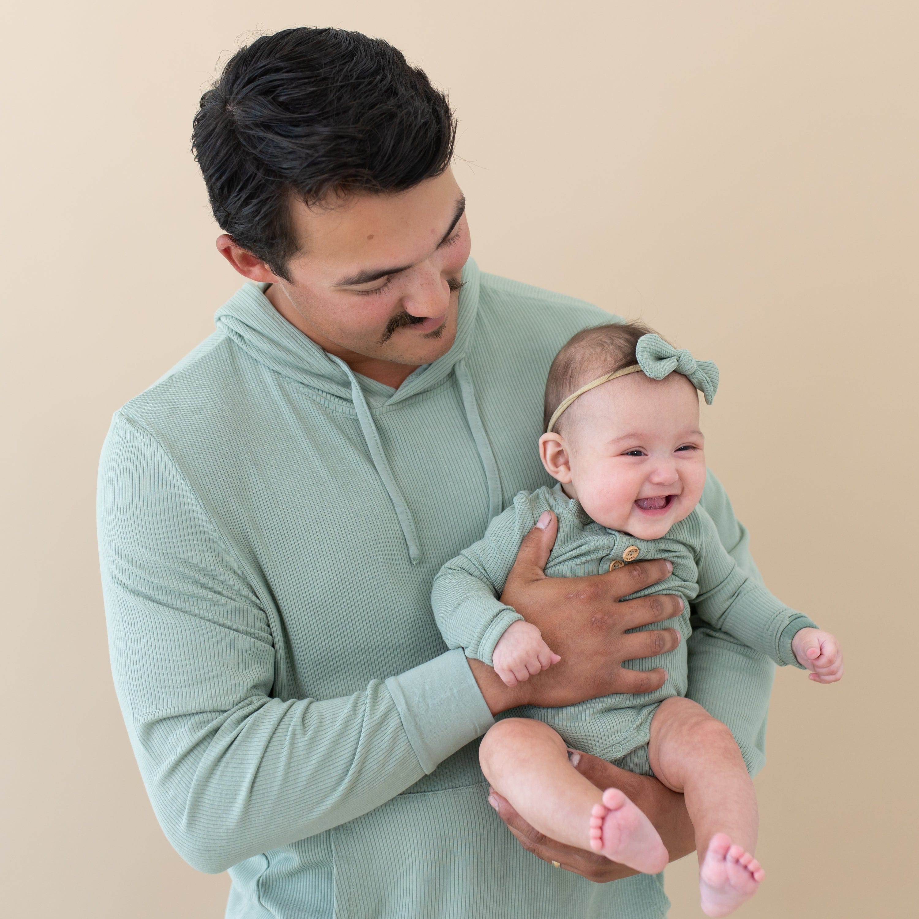 Dad wearing Kyte Baby Men's Ribbed Hoodie in Thyme holding baby in matching bodysuit