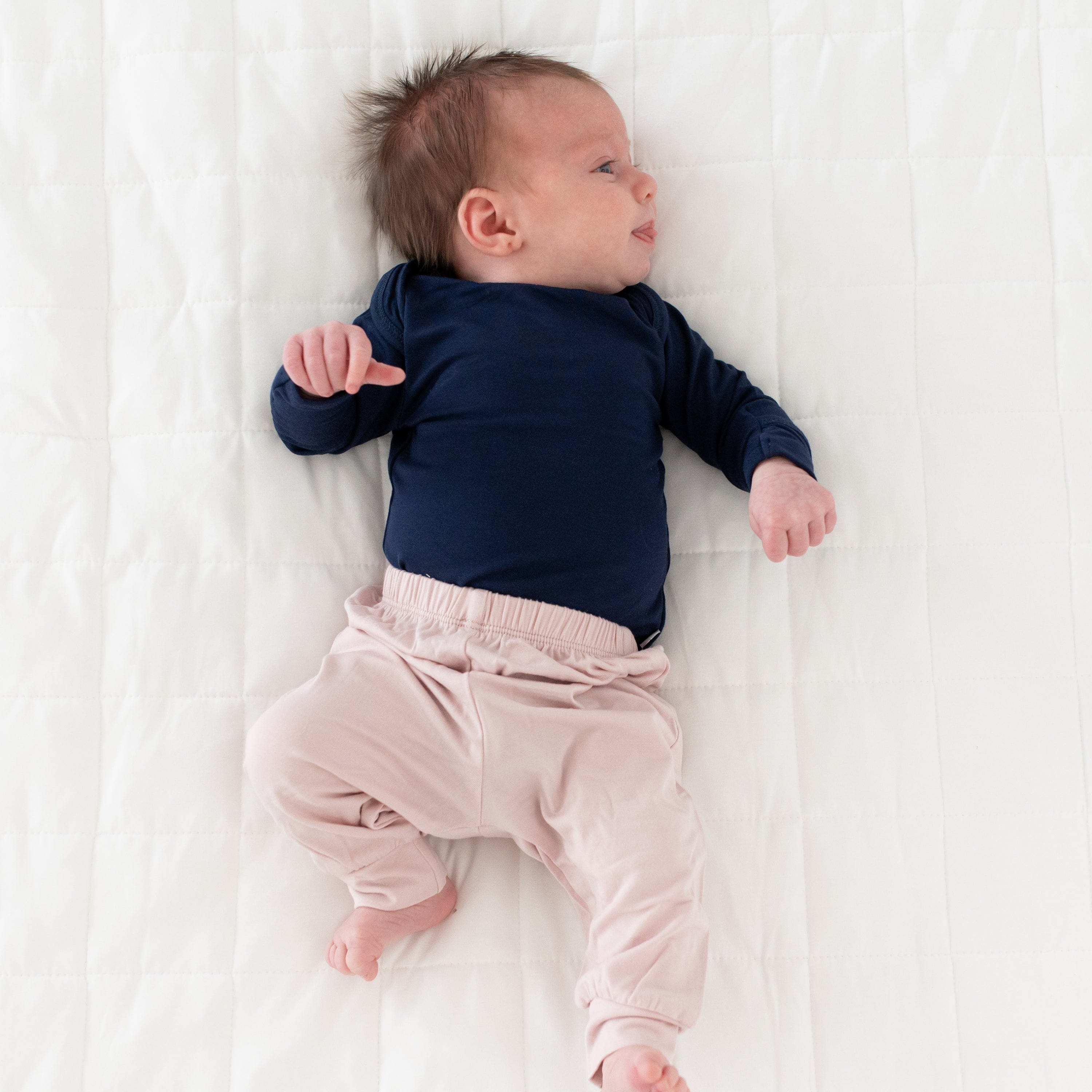 Newborn wearing Kyte Baby Pants in Blush while laying on the quilted side of a Blanket in Cloud