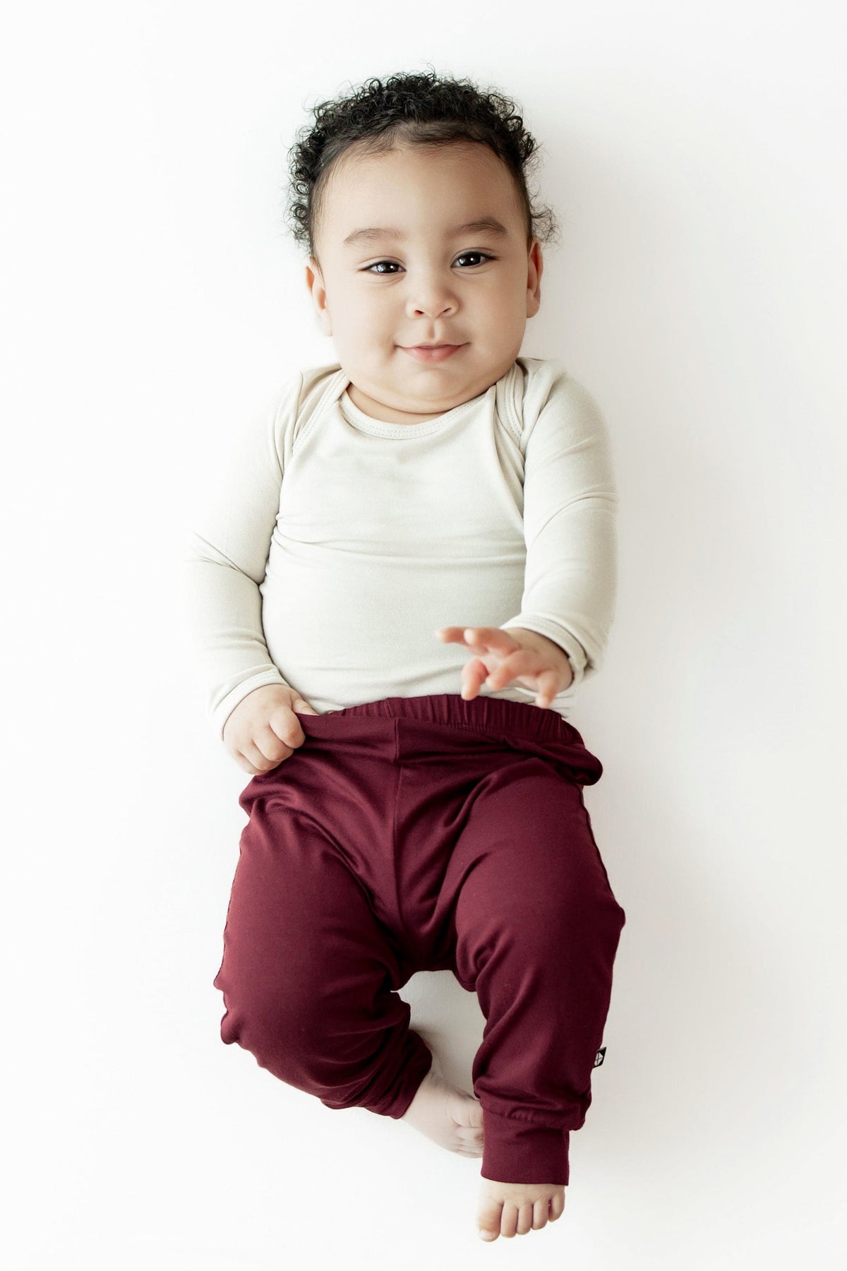A baby lying on their back against a white background, wearing a cream long-sleeve top and burgundy pants.