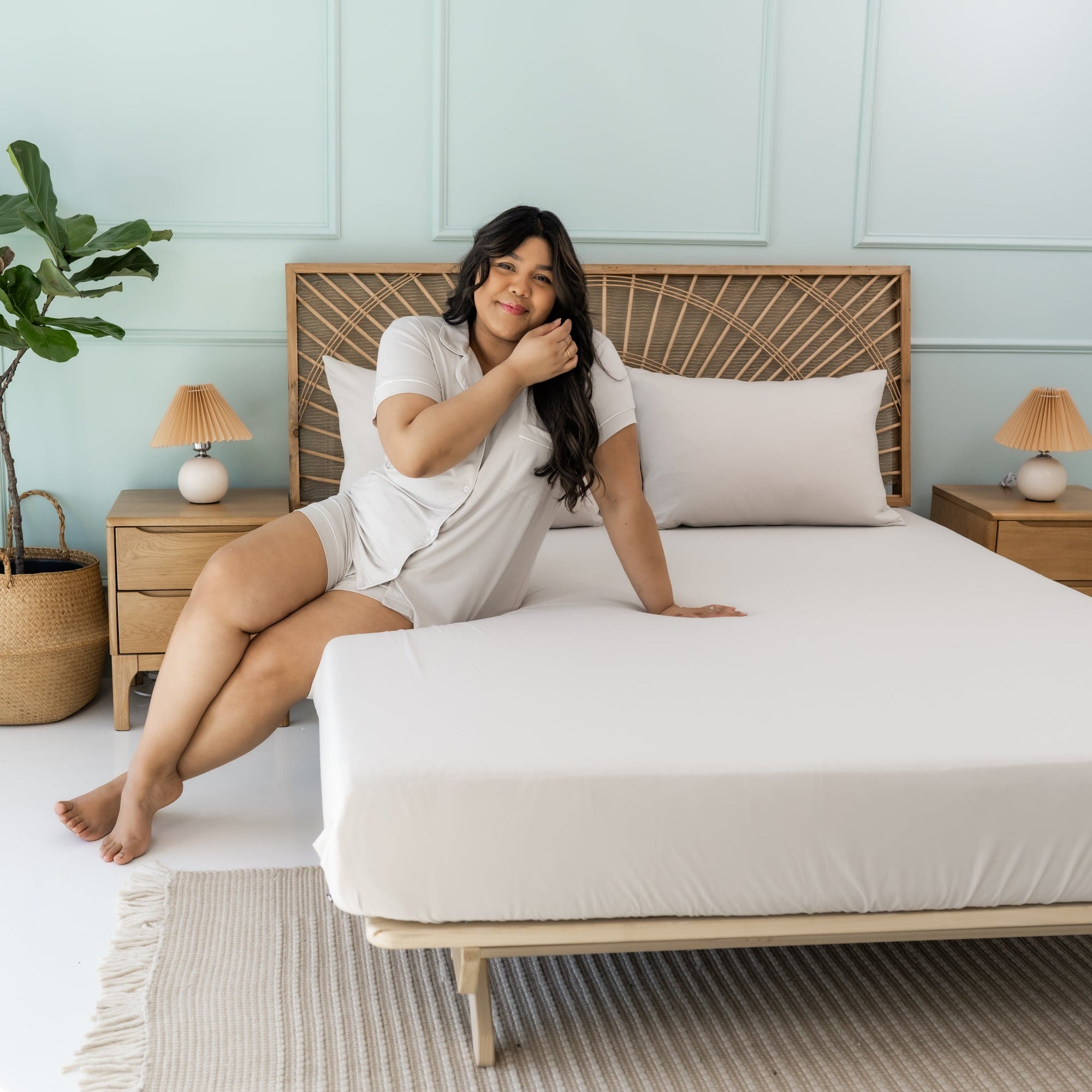 Woman sitting on bed with Kyte Baby Queen Fitted Sheet with Pillowcases in Oat