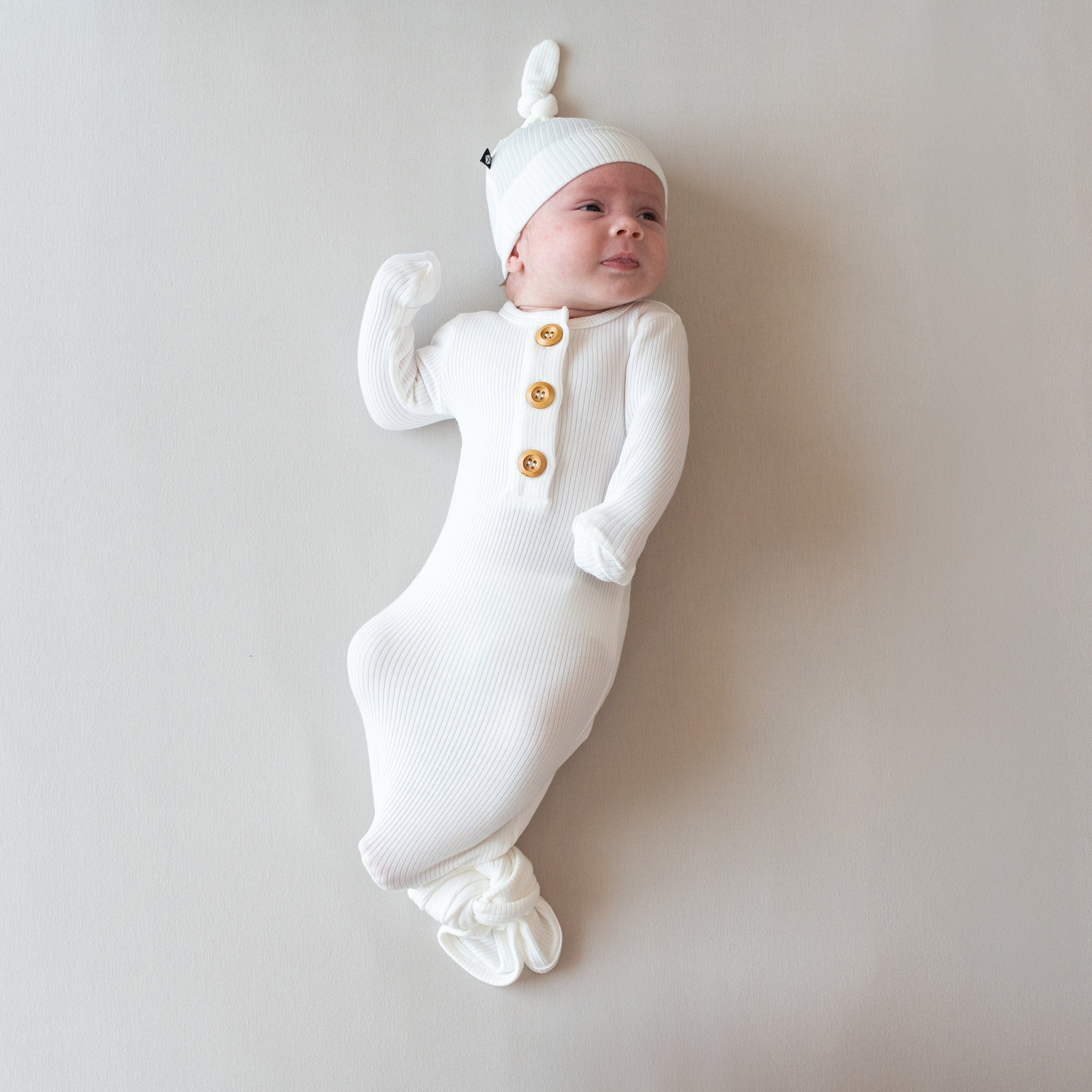 Baby laying on eggshell surface wearing Kyte Baby Ribbed Knotted Gown with Hat Set in Cloud. Baby is looking to the upper right-hand corner
