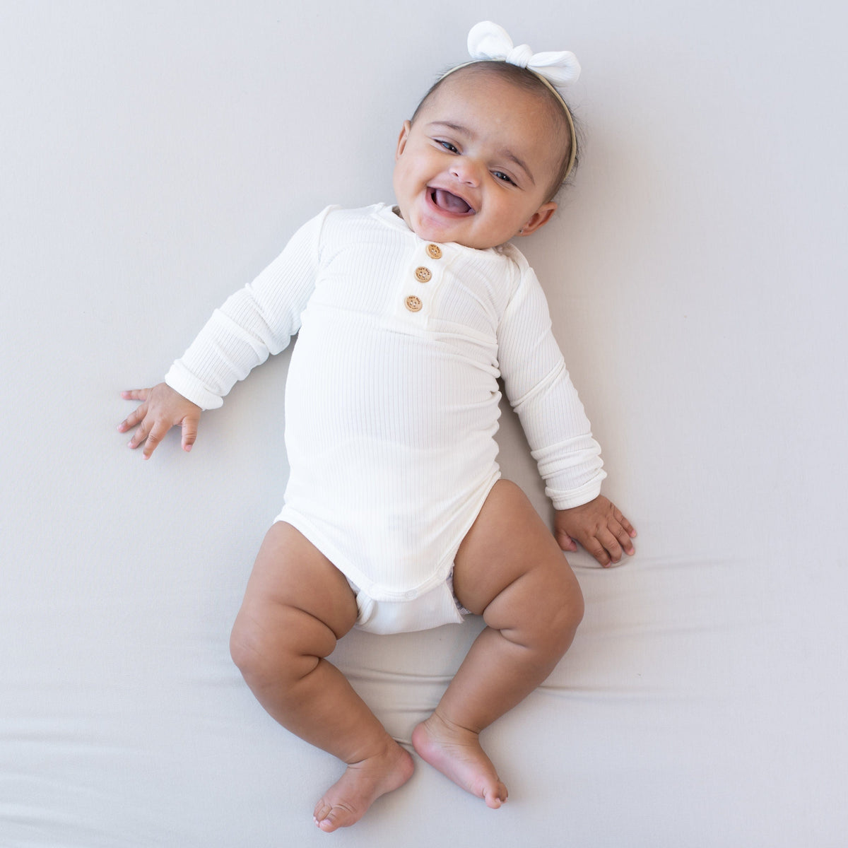 Smiling infant wearing Kyte Baby Ribbed Long Sleeve Henley Bodysuit in Cloud with a Nylon Bow in Cloud