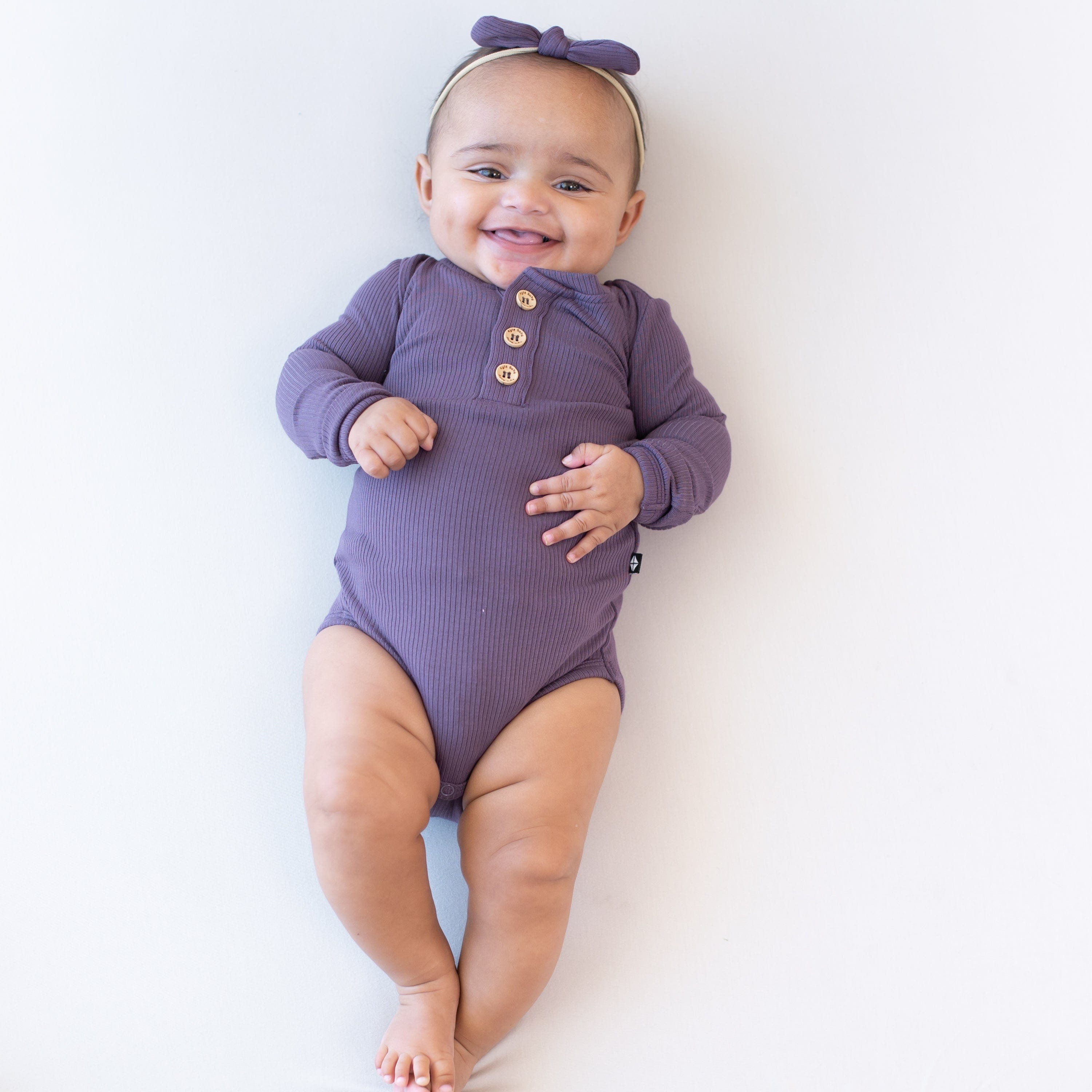 Infant wearing Kyte Baby Ribbed Long Sleeve Henley Bodysuit in Currant