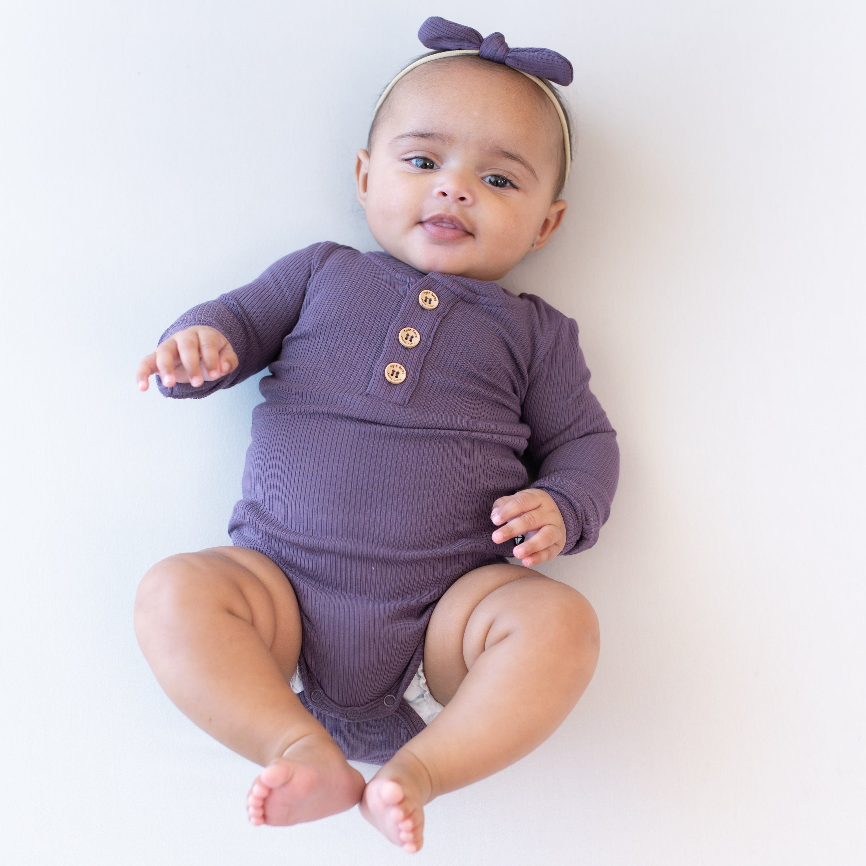 Infant wearing Kyte Baby Ribbed Long Sleeve Henley Bodysuit with snap closure in Currant