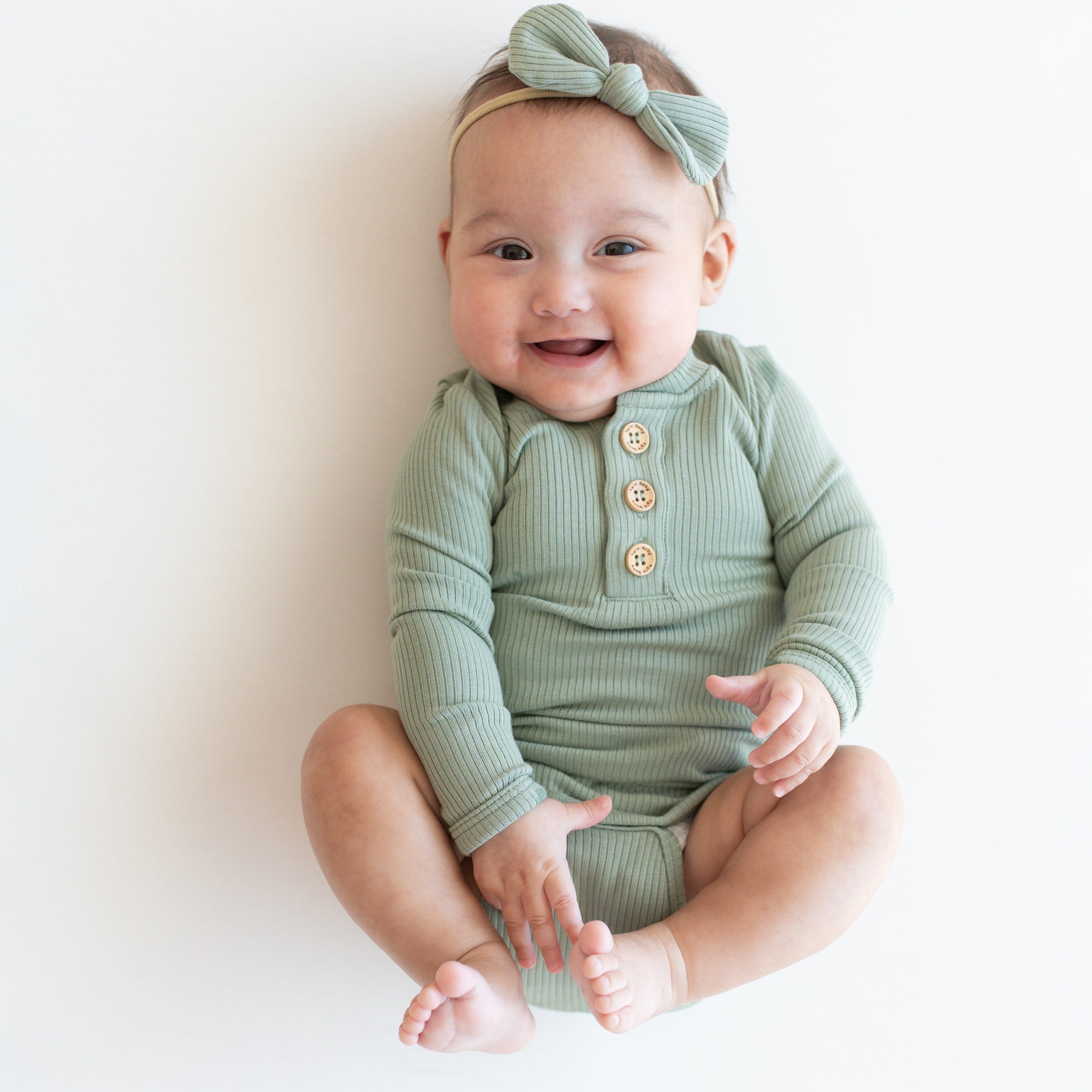 Infant wearing Kyte Baby Ribbed Long Sleeve Henley Bodysuit with snap closure in Thyme with matching Nylon Bow