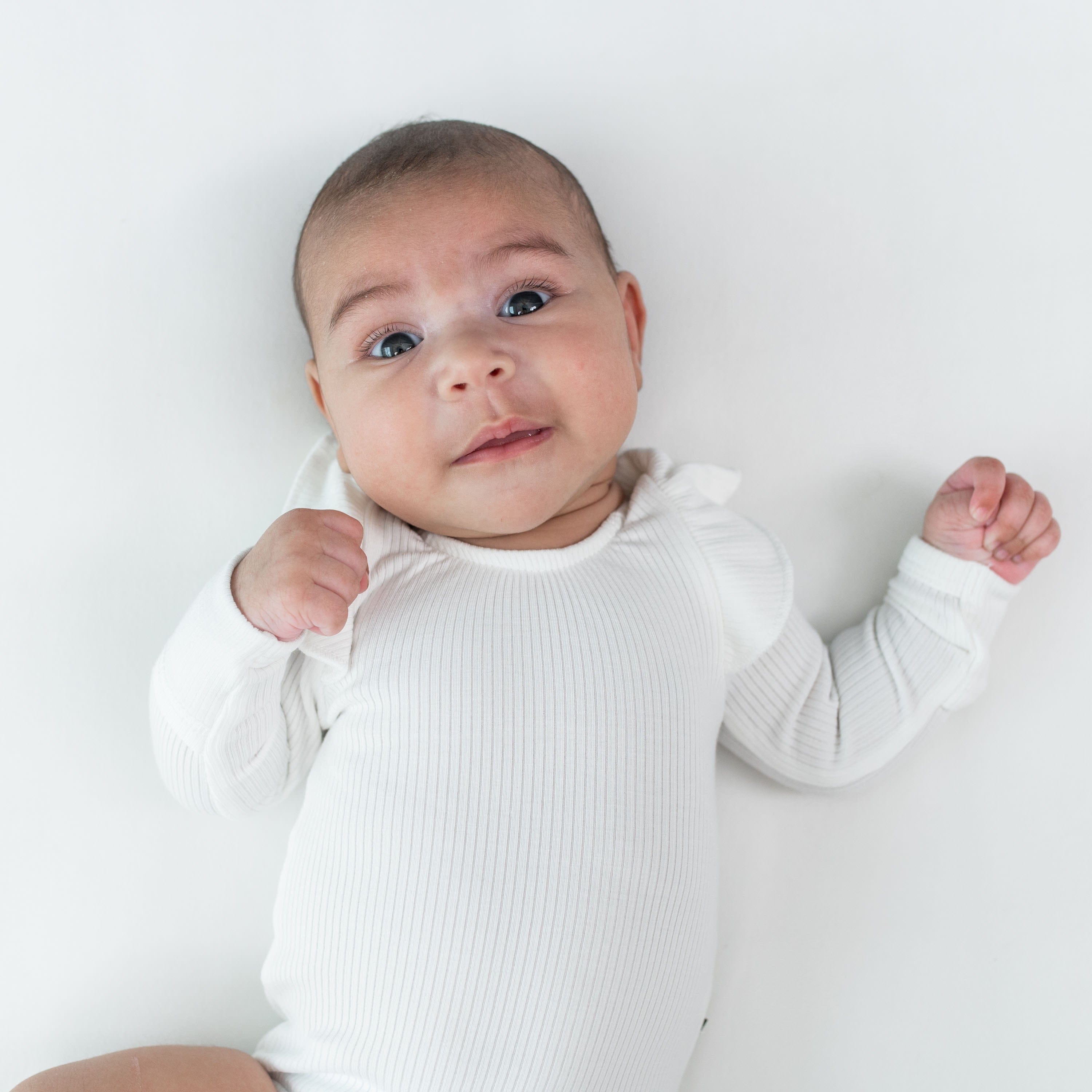 Close up of infant laying on white surface and wearing Kyte Baby Ribbed Long Sleeve Ruffle Leotard in Cloud.