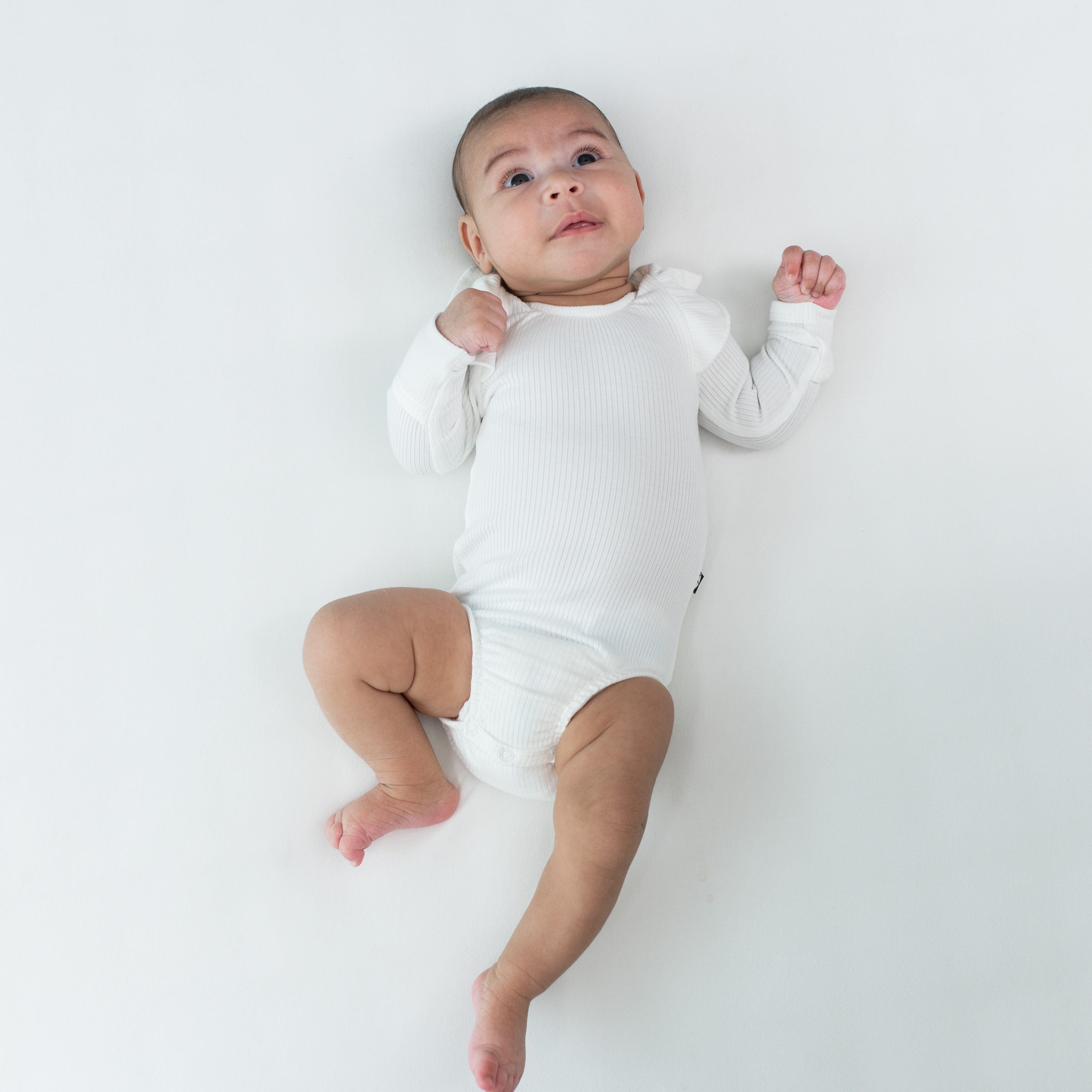 Baby laying on white surface wearing Kyte Baby Ribbed Long Sleeve Ruffle Leotard in Cloud. Baby is looking upwards