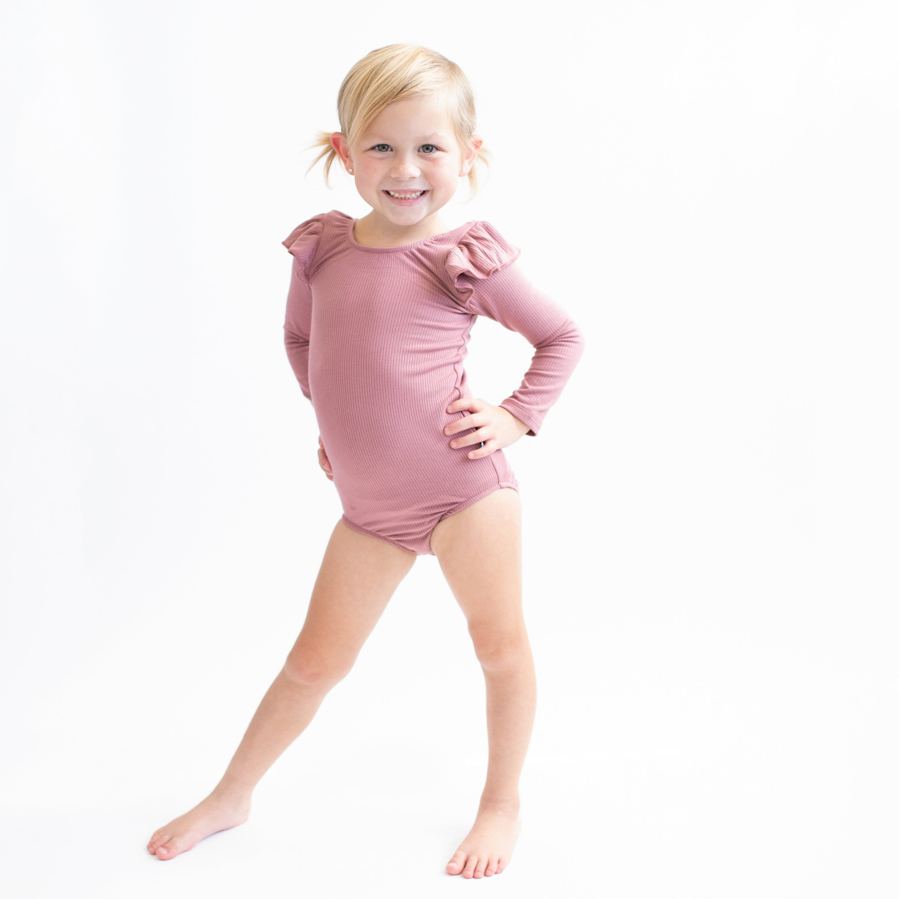Girl wearing Kyte Baby bamboo Ribbed Long Sleeve Leotard in Dusty Rose with Shoulder Ruffle Detail