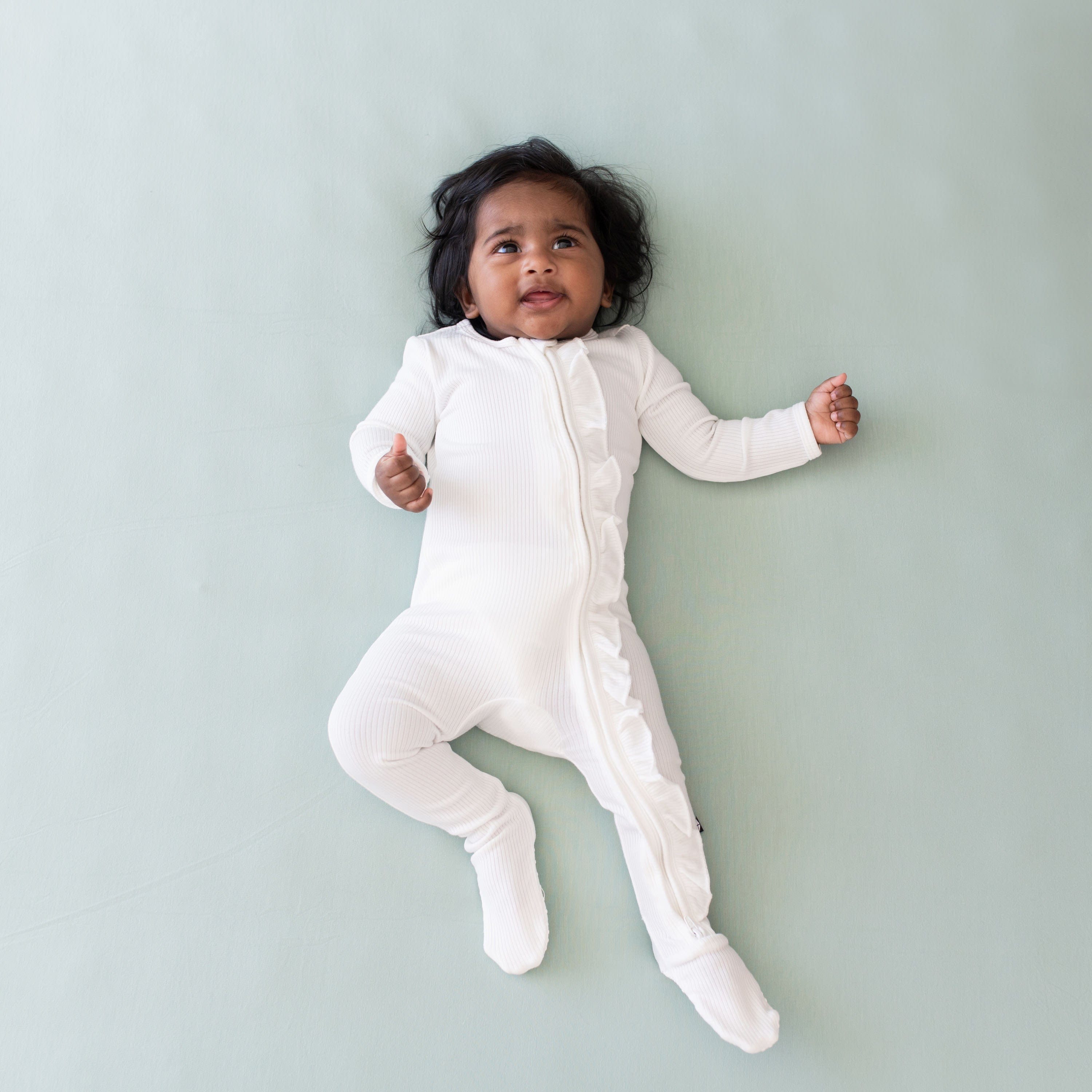 Infant laying on sage background, looking upwards and wearing Kyte Baby Ribbed Zipper Footie with Ruffle in Cloud