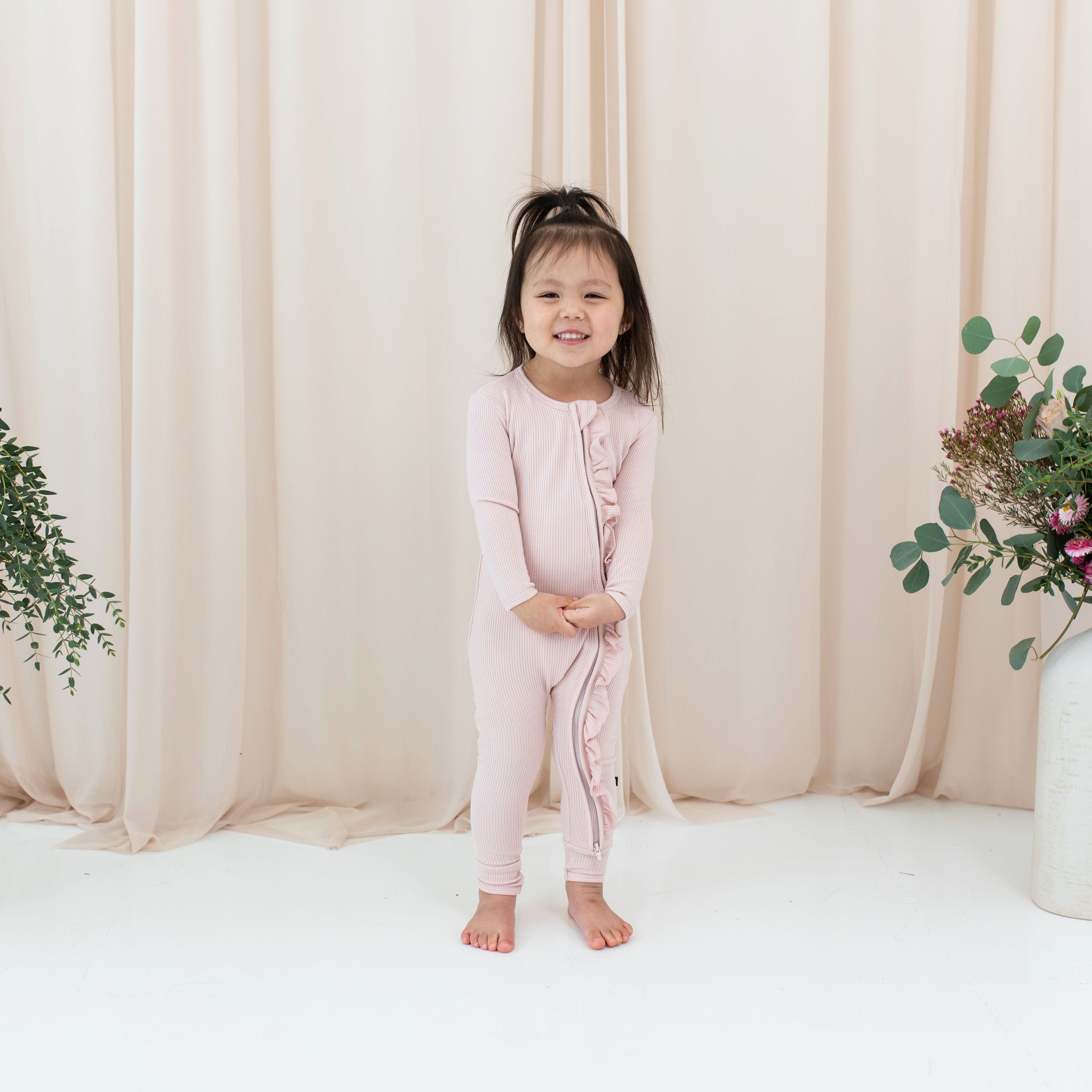 Girl wearing Kyte Baby bamboo Ribbed Ruffle Zipper Romper in Blush