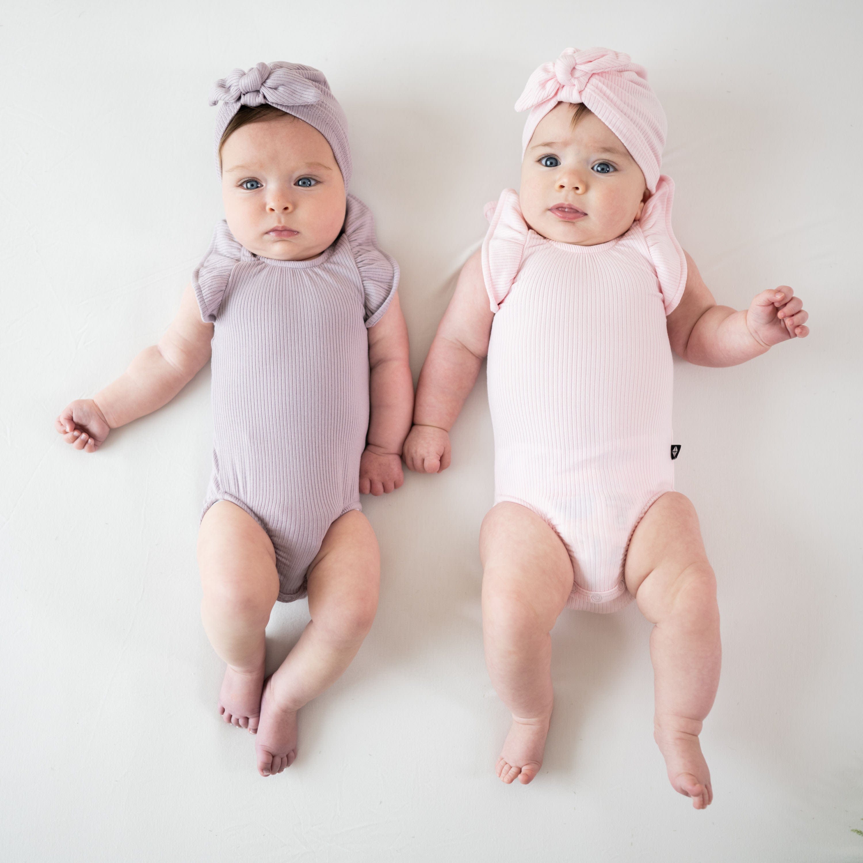 Babies wearing Kyte Baby Ribbed Sleeveless Ruffle Leotards with matching bow hats