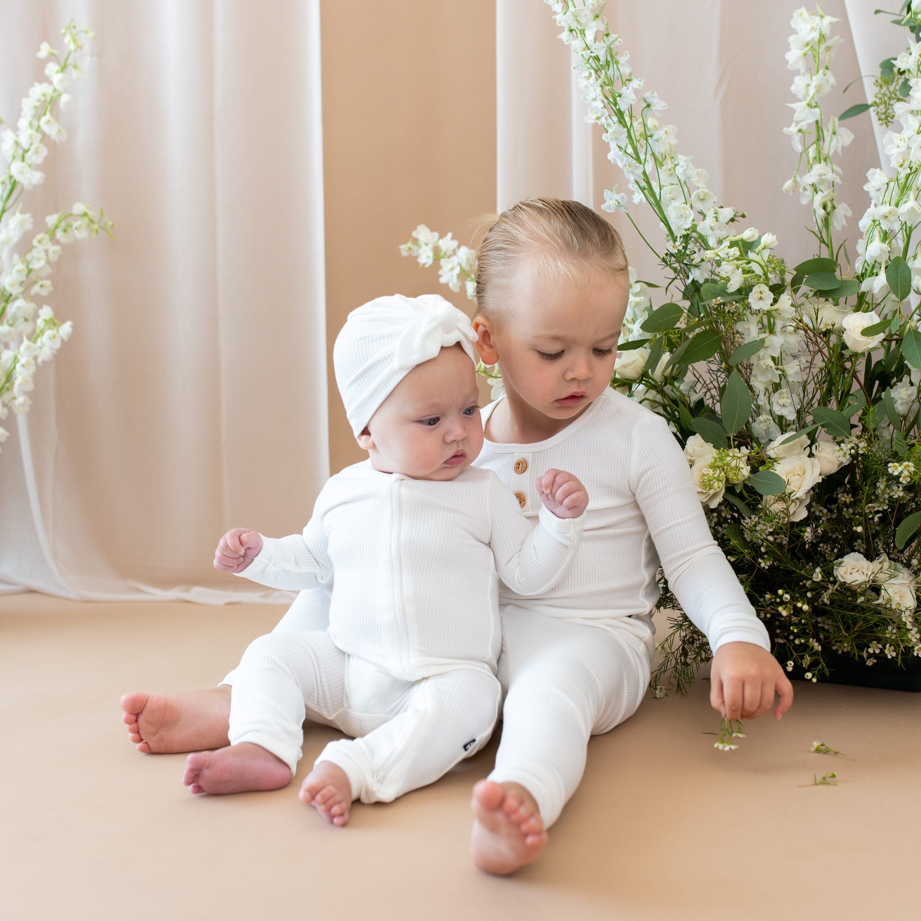 Siblings sitting next to a floral bush in matching Kyte Baby bamboo outfits in Cloud