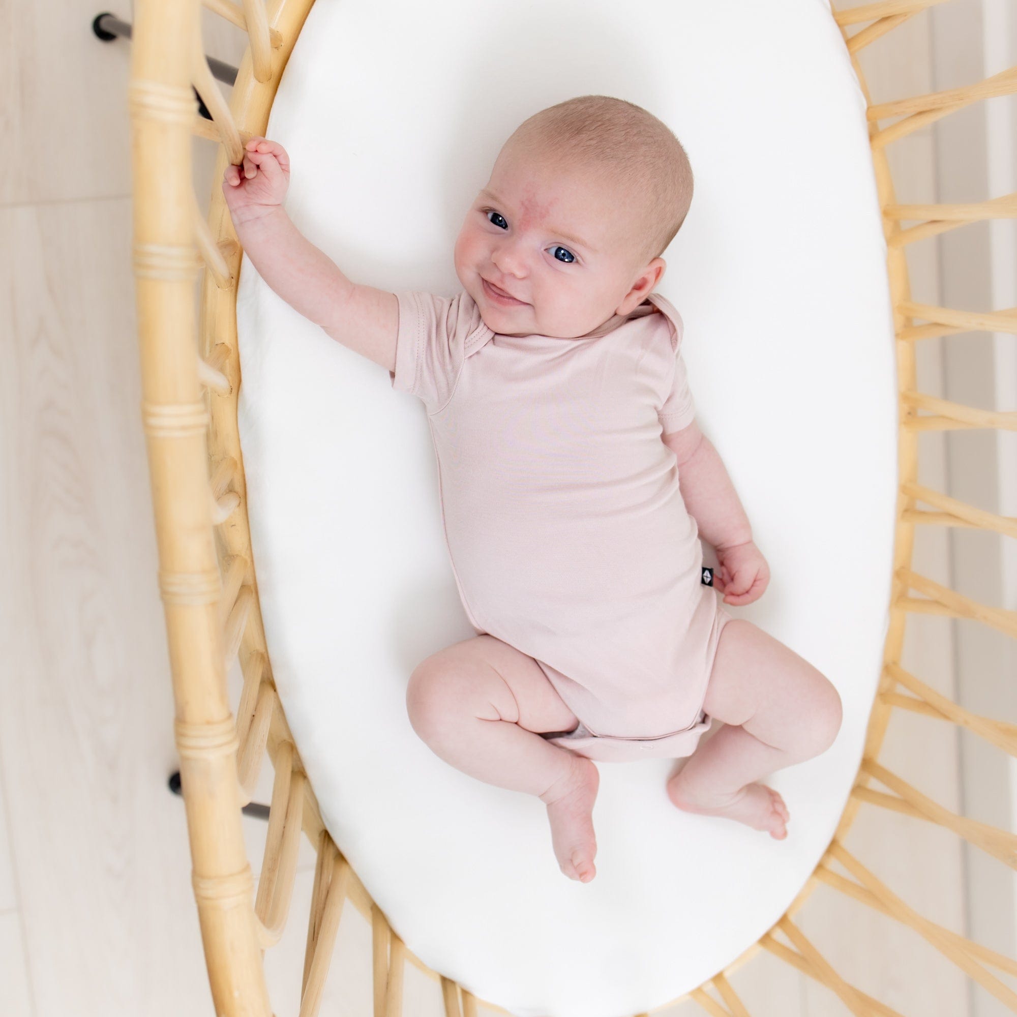 Smiling infant laying in rattan bassinet while wearing Kyte Baby Short Sleeve Bodysuits Bodysuit in Blush