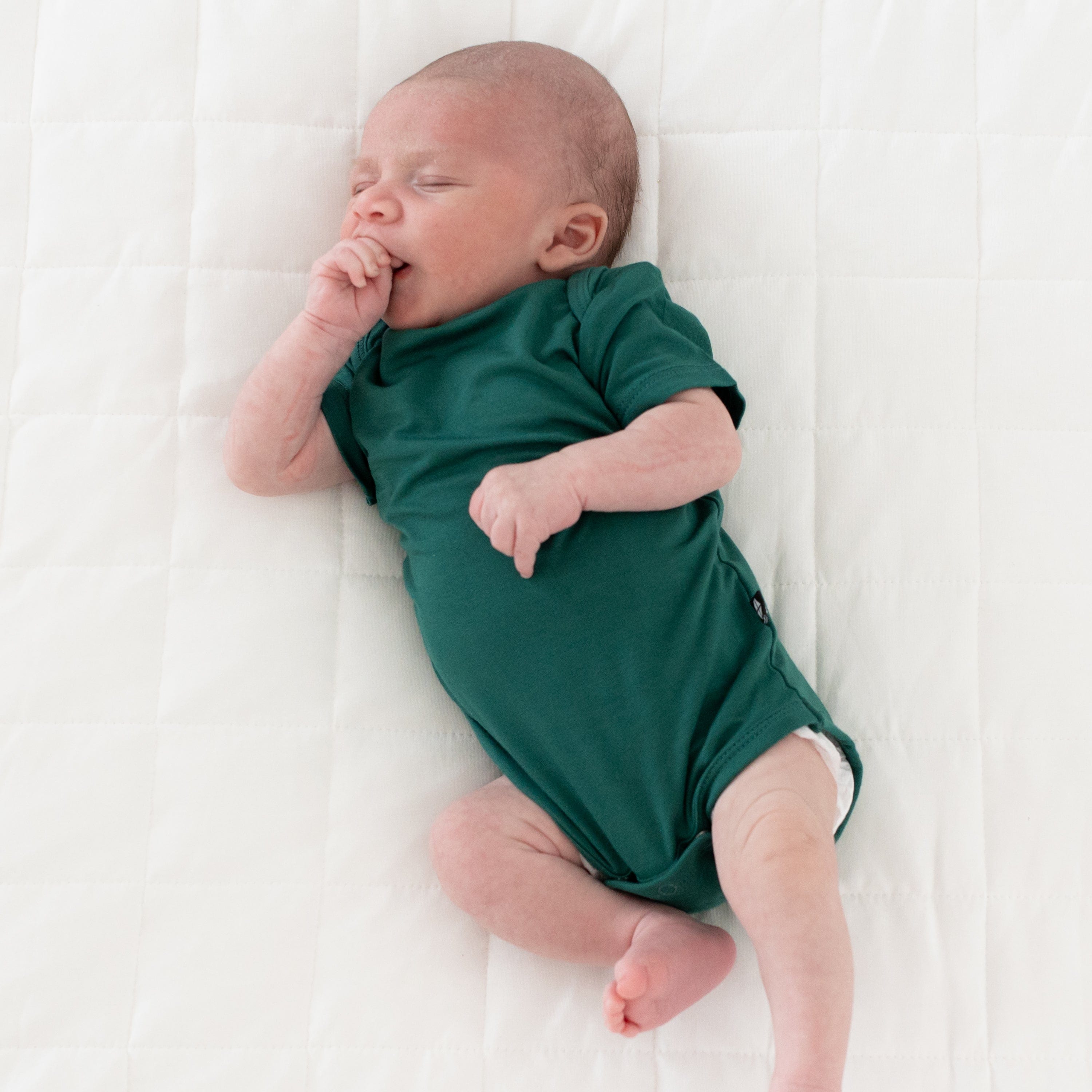 Sleeping newborn wearing Kyte Baby Bodysuit in Emerald with hand brought up to mouth