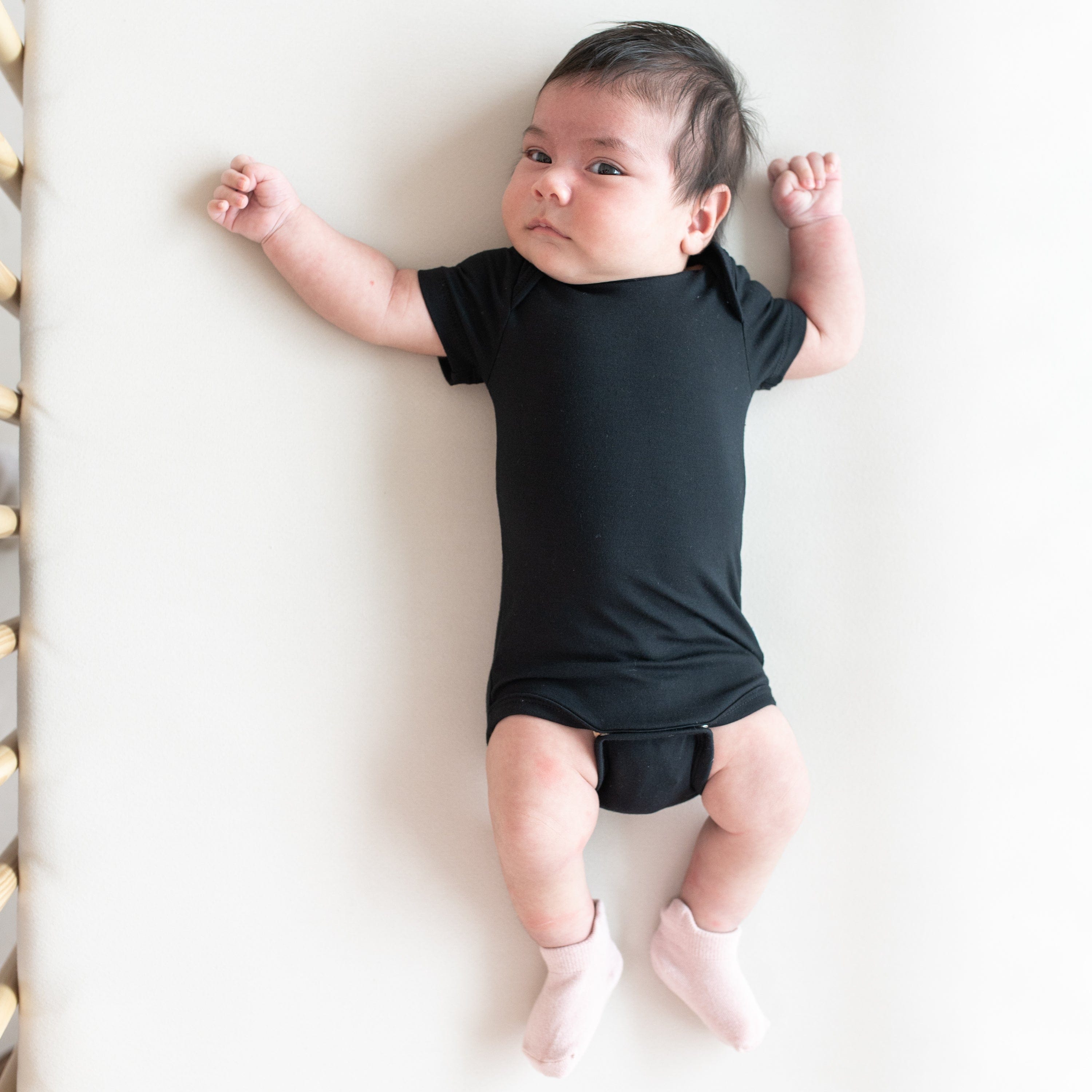 Baby wearing Kyte Baby infant Bodysuit in Midnight while laying on a Crib Sheet in Oat