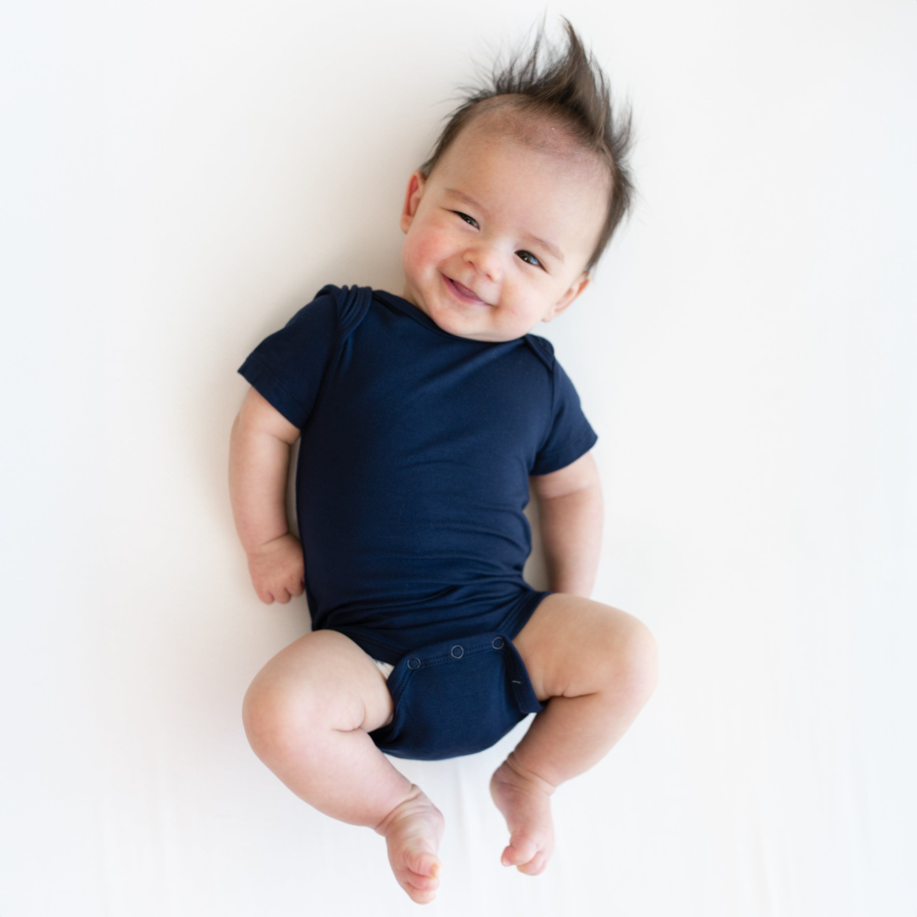 Smiling baby laying on white surface while wearking Kyte Baby Bodysuit Bodysuit in Navy