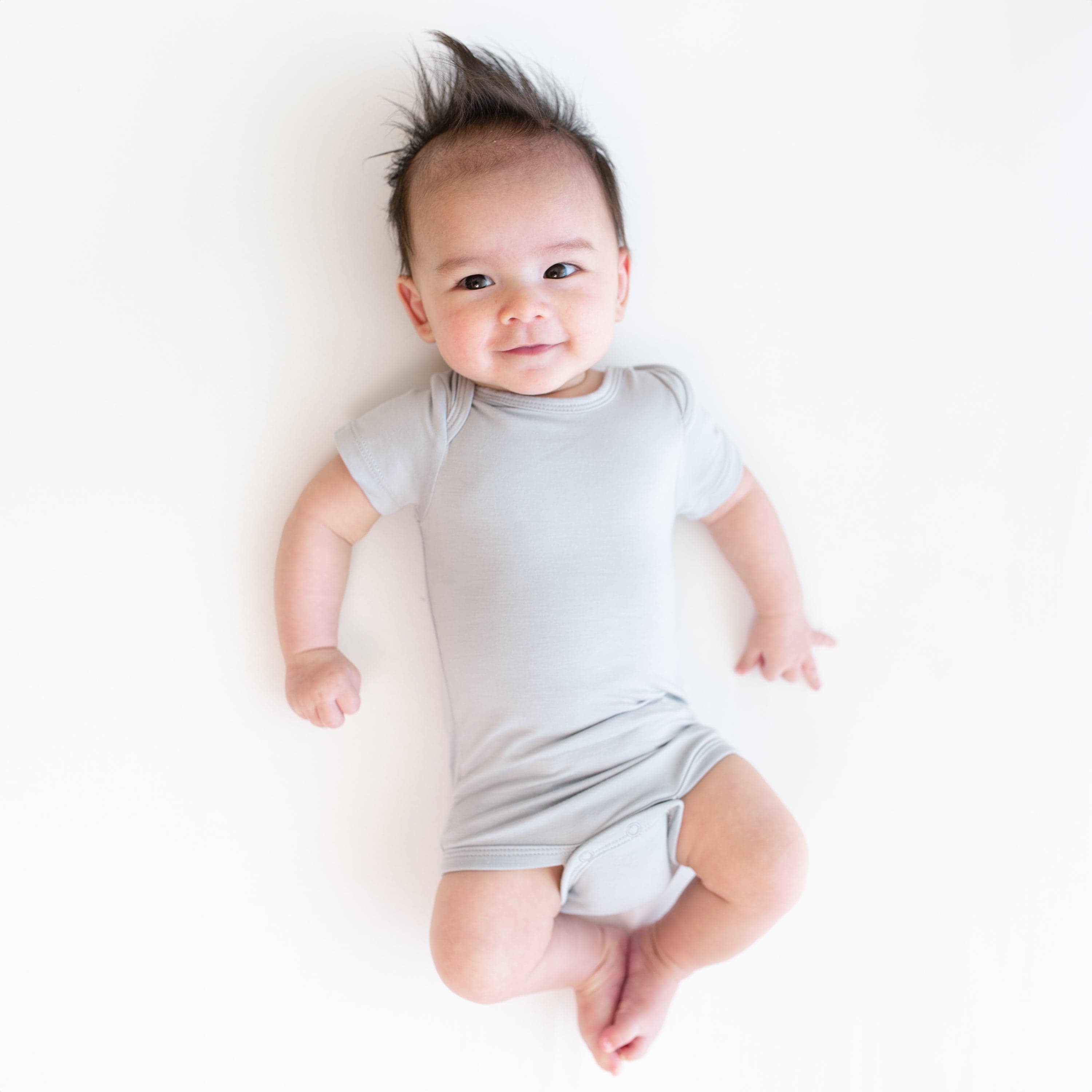 Smiling baby laying on a white surface and wearing Kyte Baby Bodysuit in Storm