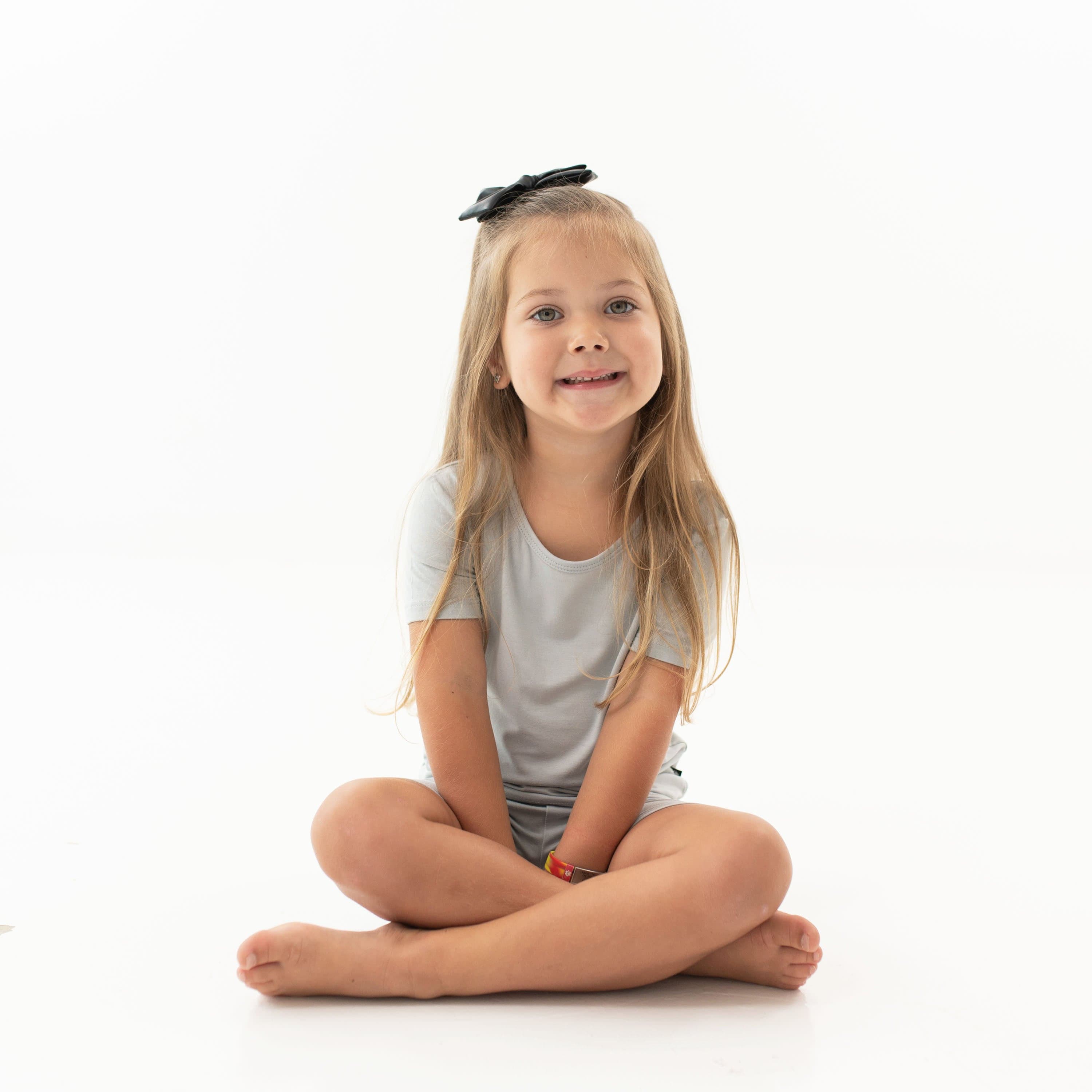Child sitting cross-legged while wearing the Short Sleeve Pajamas in Storm with a Midnight bow in her hair