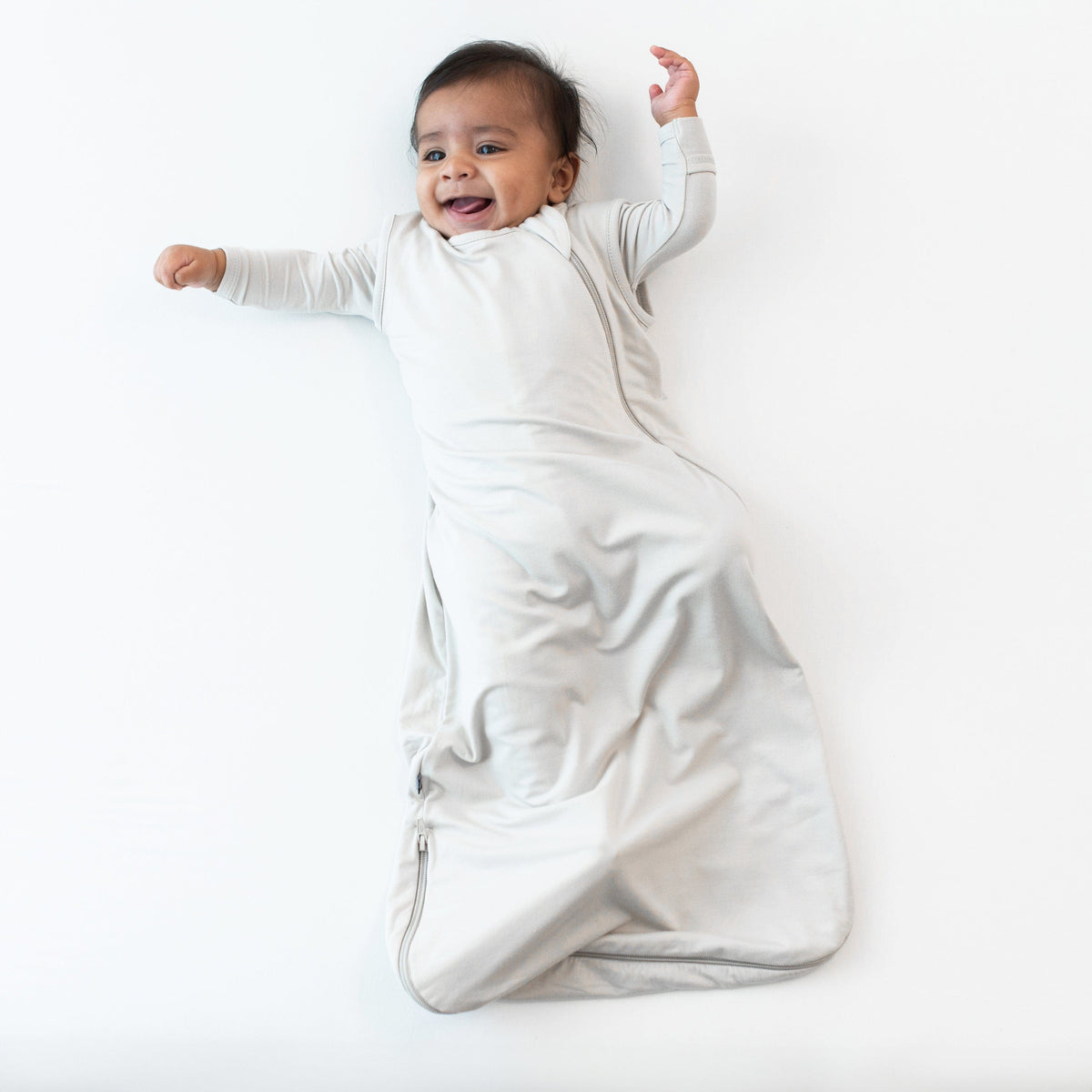 Smiling infant wearing the 0.5 Tog Sleep Bag in Oat with an Oat long sleeve romper underneath