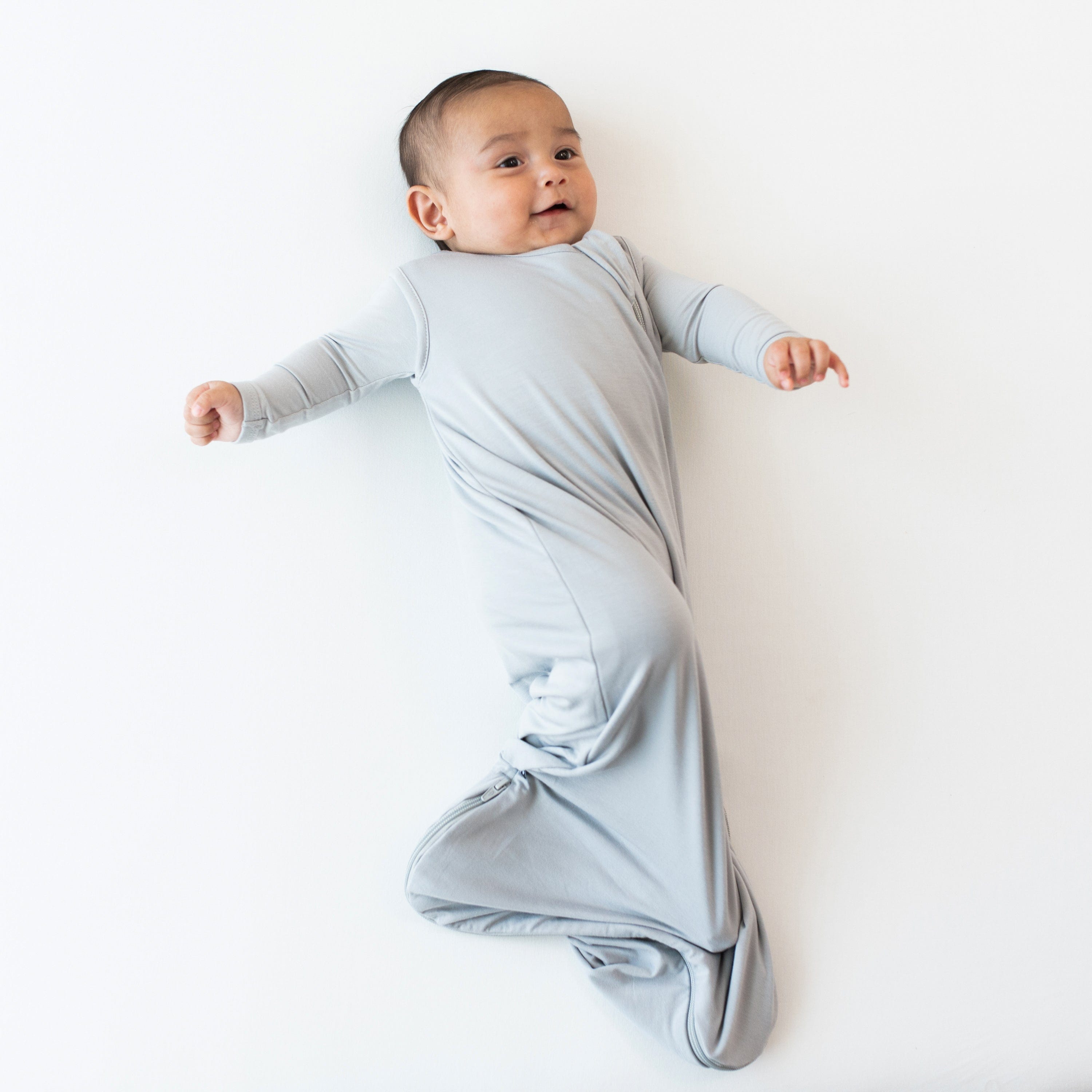 Infant laying on a white surface while wearing the 0.5 Tog Sleep Bag in Storm with a matching long sleeve bodysuit underneath