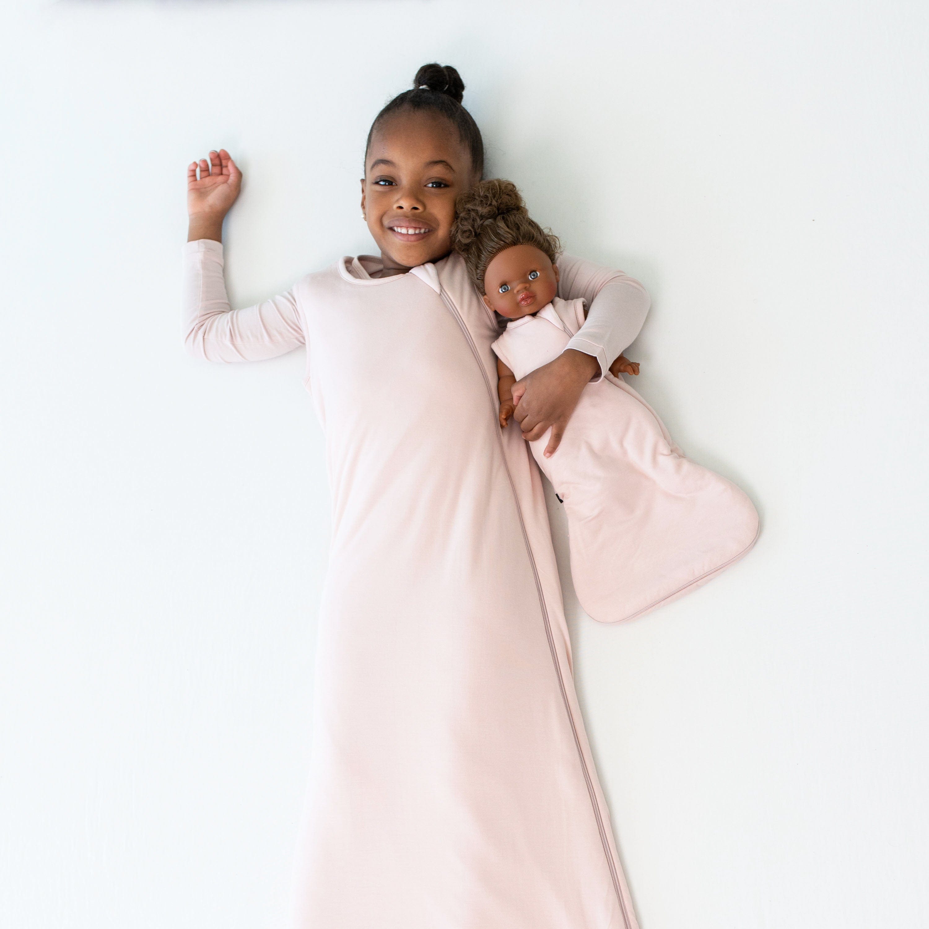 Young child wearing the Sleep Bag in Blush 1.0 holding a doll that is in a matching doll sleep bag