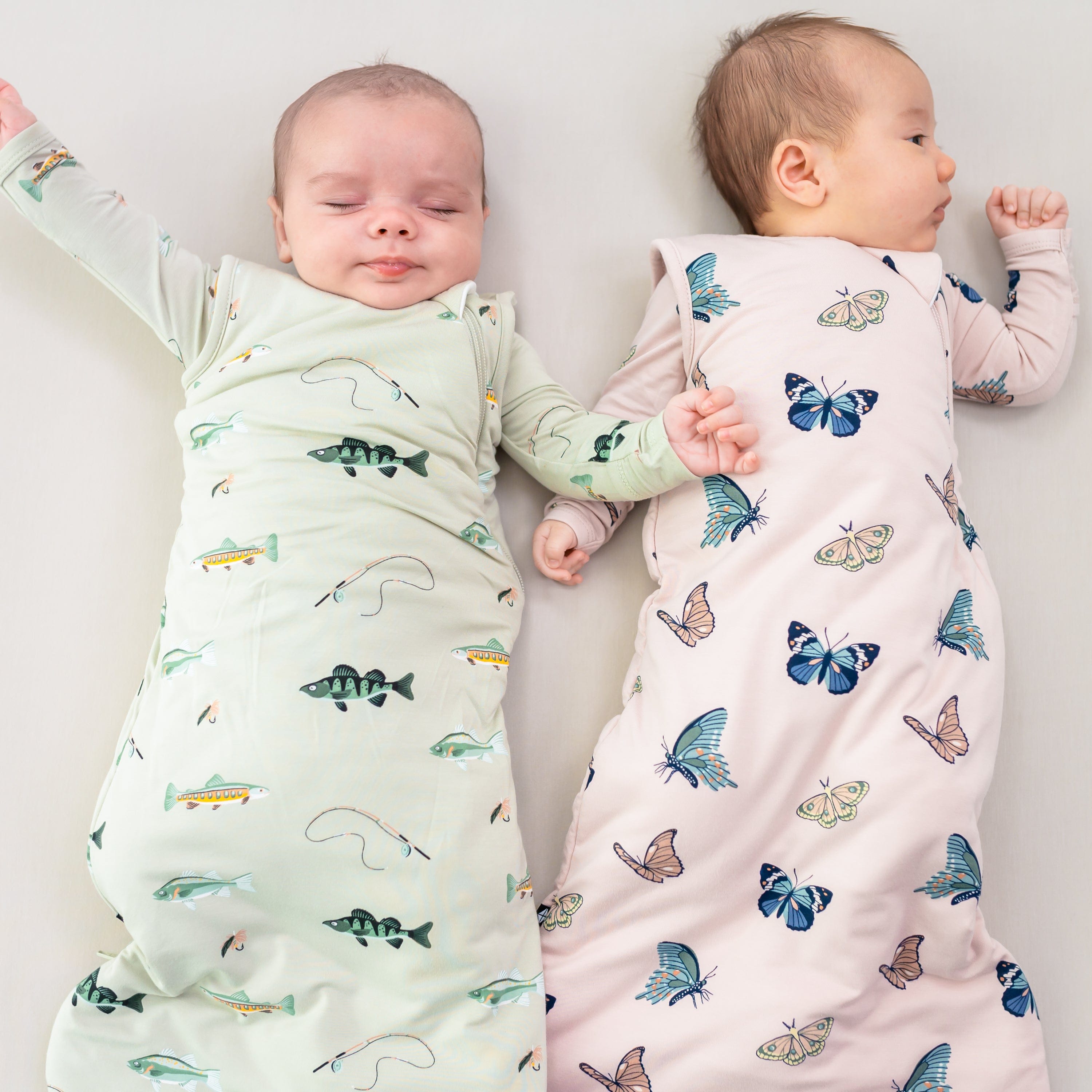 Two infants wearing 1.0 Sleep Bags in Fishing and Blush Butterfly