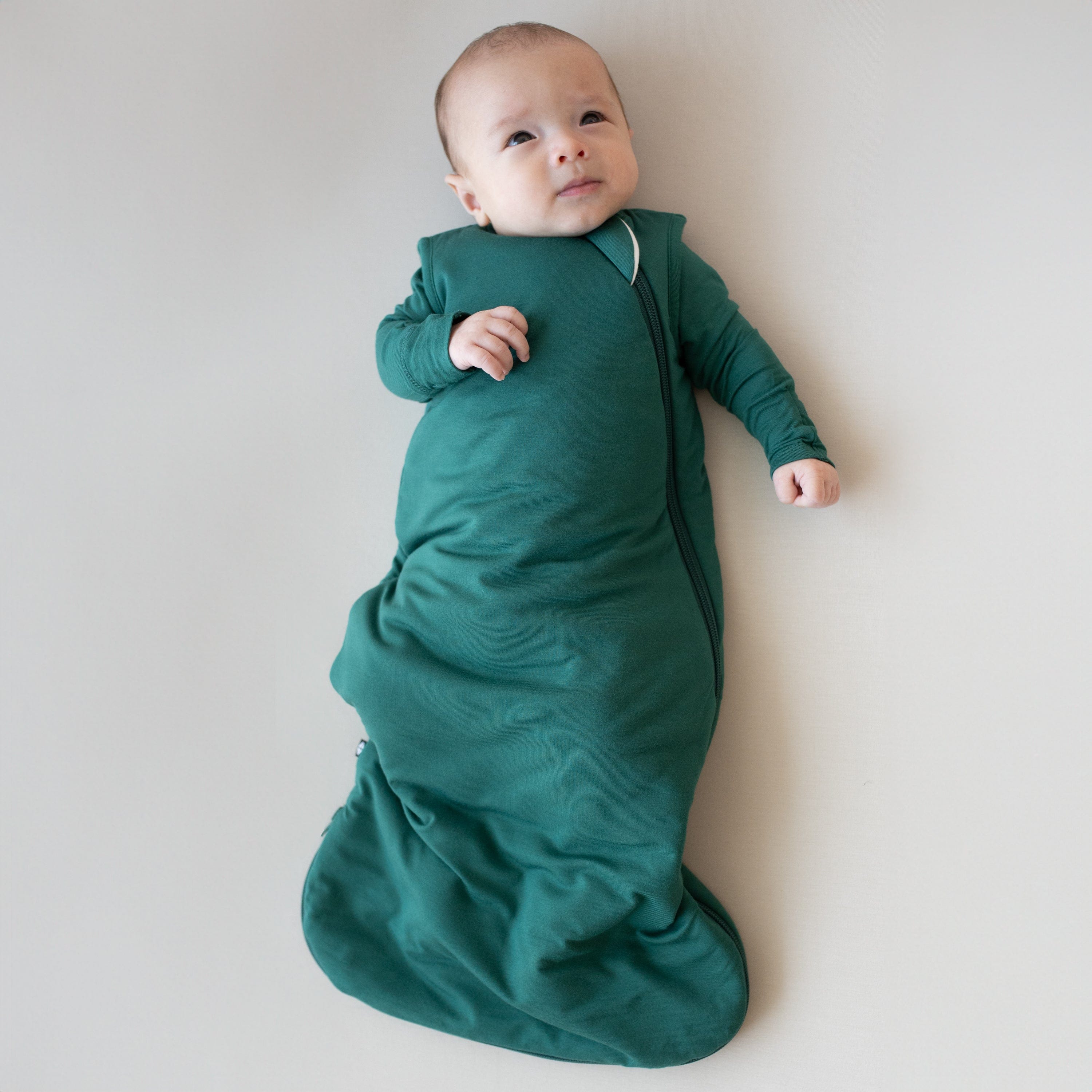 Baby laying on an off-white surface while looking upwards. Baby is wearing the Sleep Bag in Emerald 1.0
