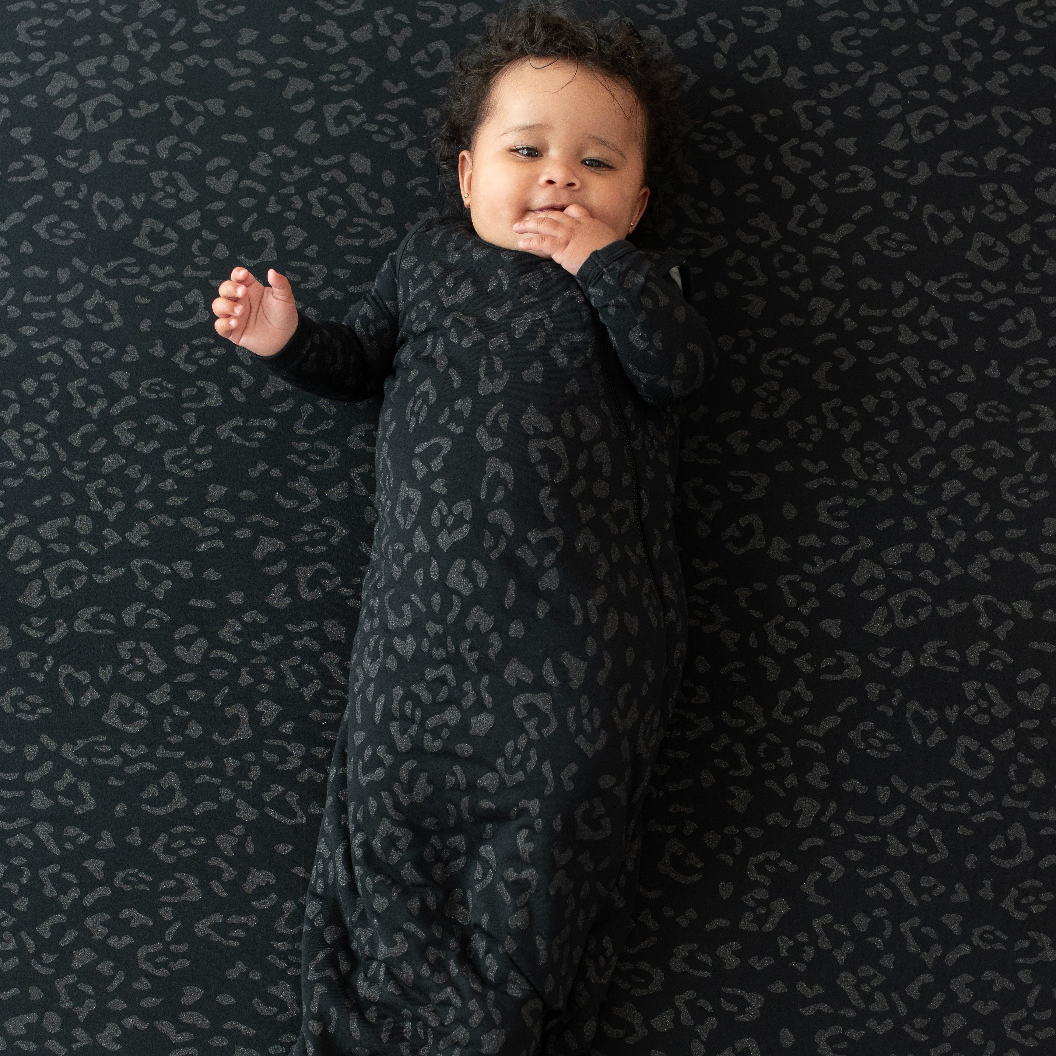 Infant modeling the Sleep Bag in Midnight Leopard 1.0 wearing a Midnight Leopard long sleeve romper laying on a Midnight Leopard blanket