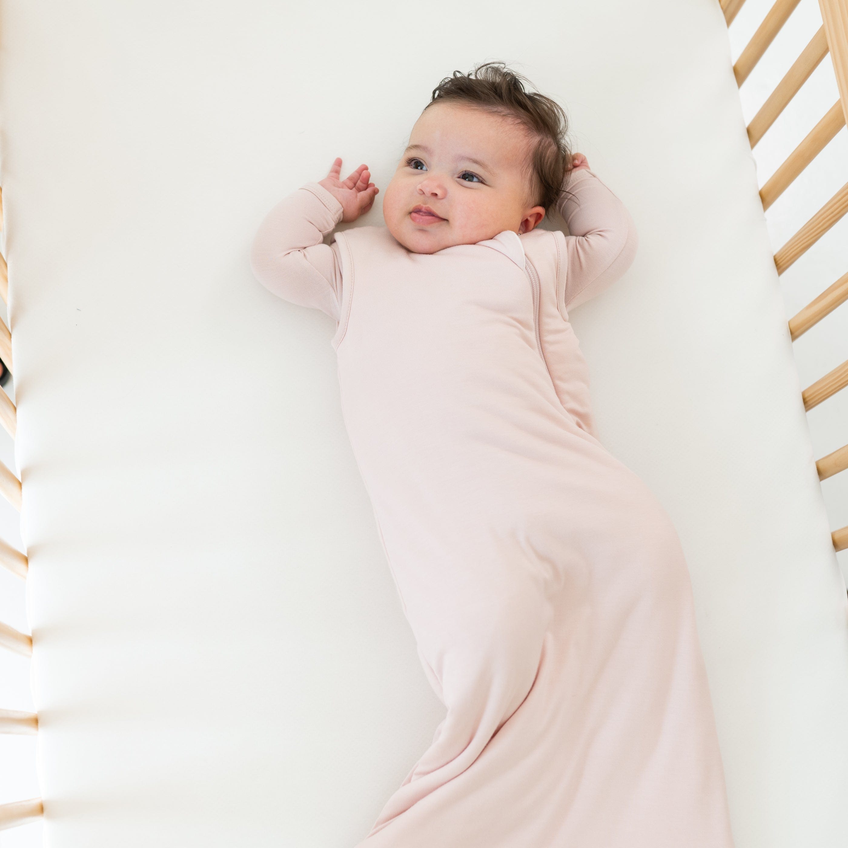 Infant laying in a wooden crib wearing a Sleep Bag in Blush 2.5 while laying on a Cloud crib sheet