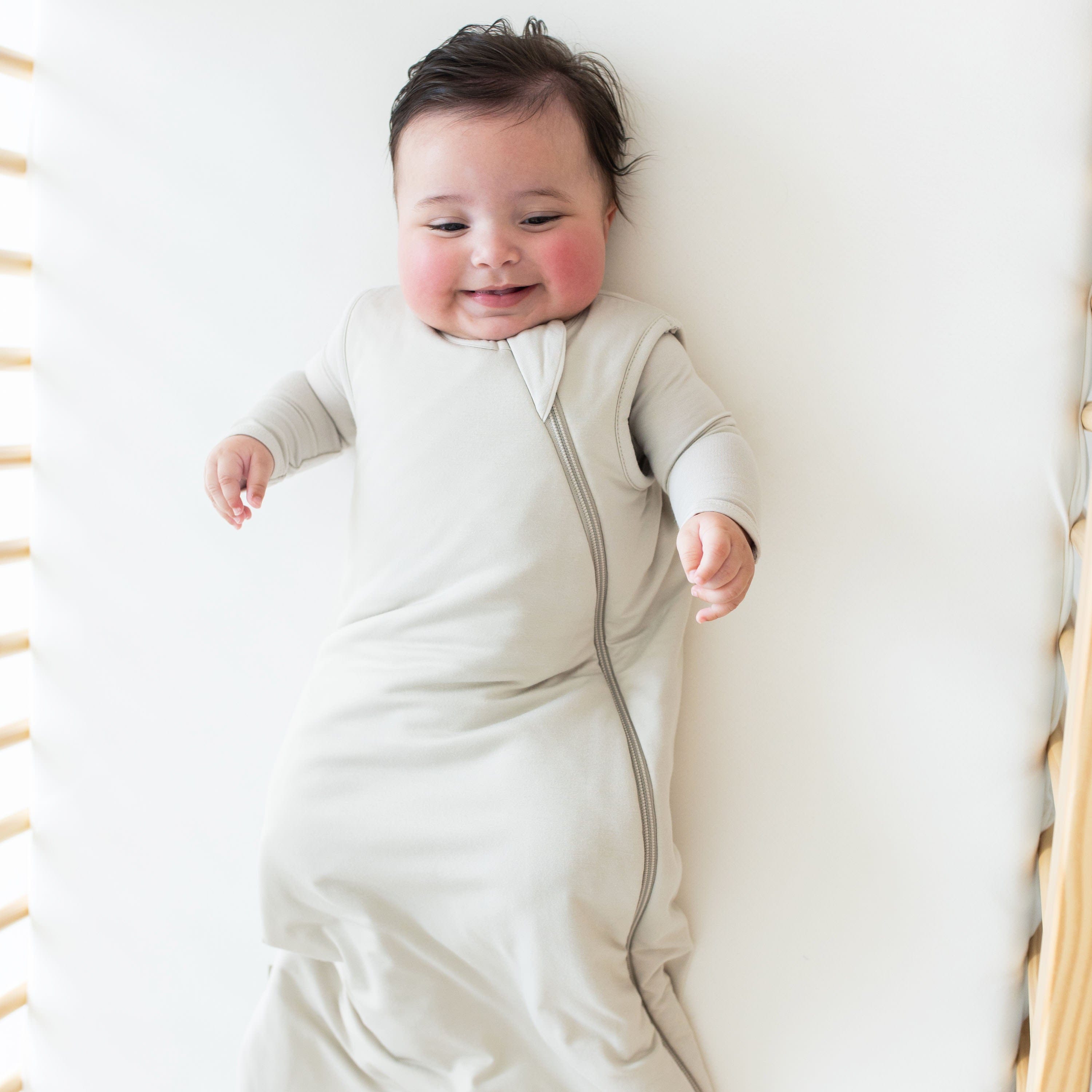 Smiling baby laying in a crib wearing Kyte Baby Sleep Bag in Oat 2.5