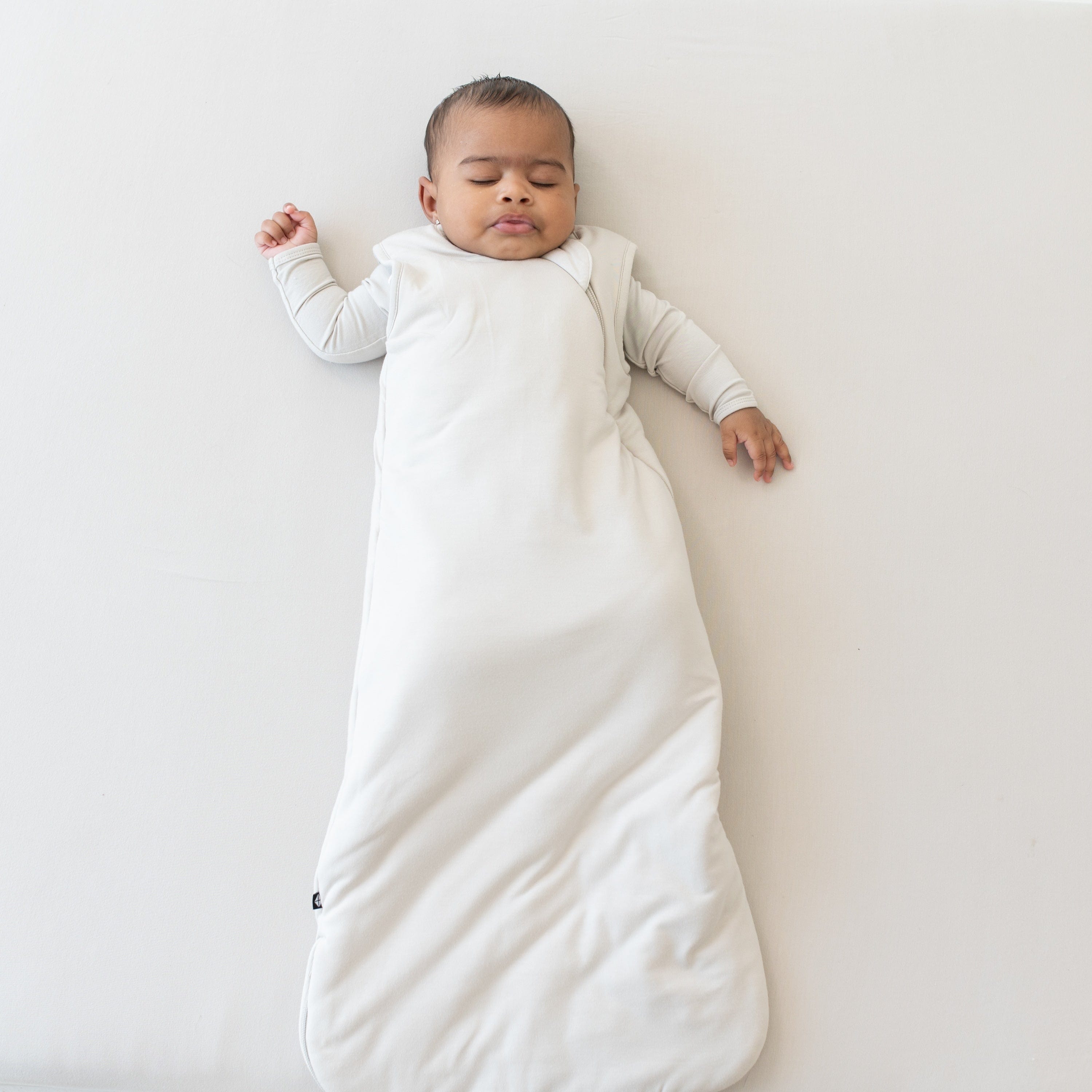 Sleeping baby wearing the Sleep Bag in Oat 2.5 with an Oat long sleeve romper underneath