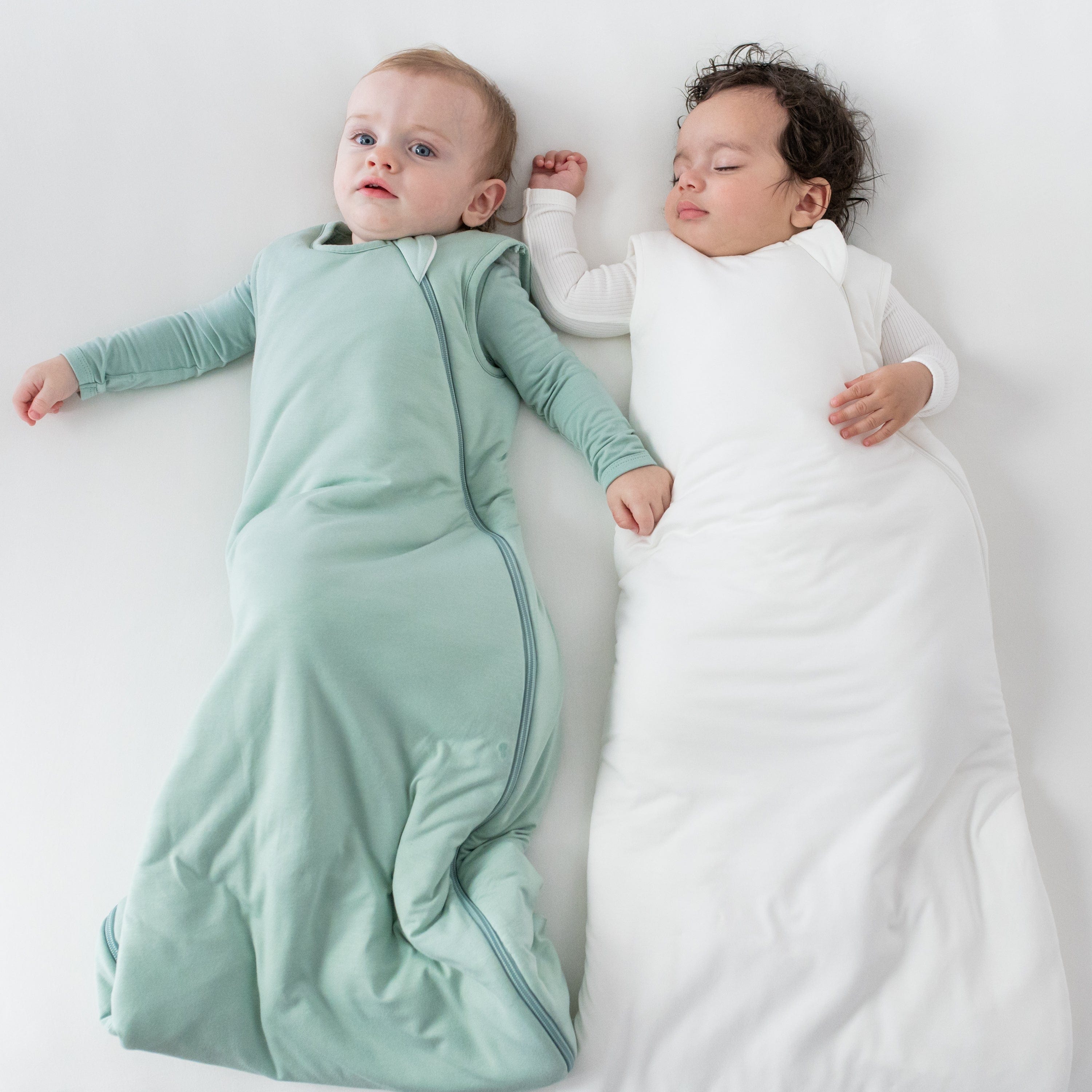 Babies laying next to each other wearing Kyte Baby sleep bags TOG 2.5 in the colors Sage and Cloud
