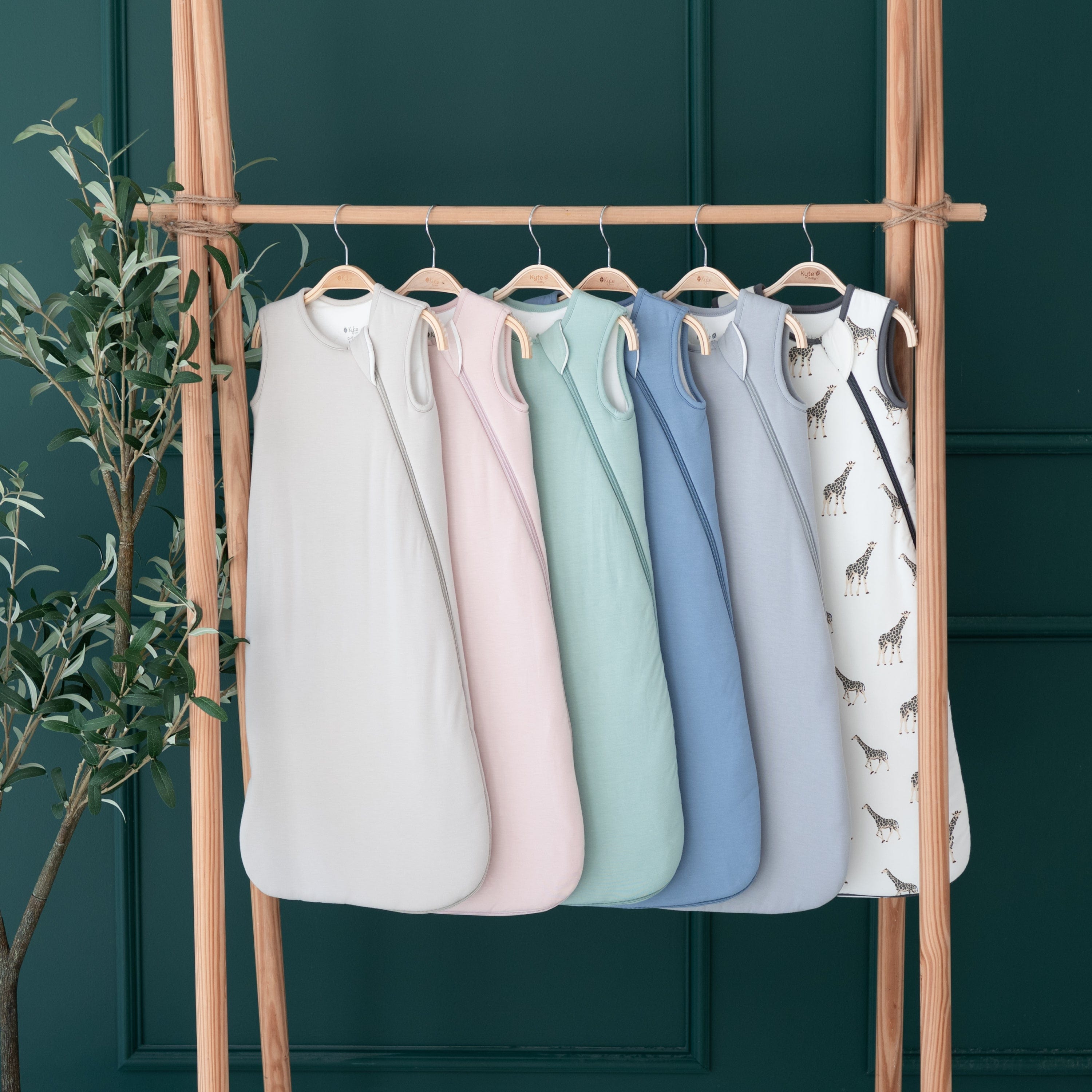 Kyte Baby 2.5 TOG Sleep Bags in different colors hanging on a wooden rack in front of an emerald green wall