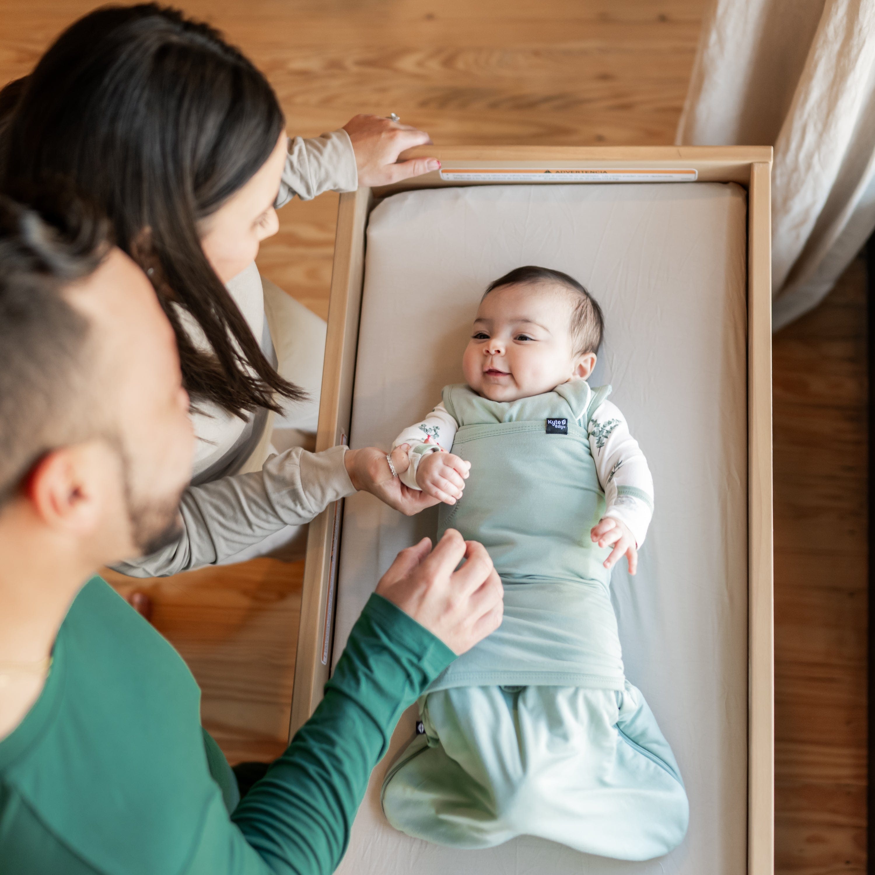 Baby in bassinet while wearing Sleep Bag Swaddler in Sage. Parents are standing next to bassinet, looking down at baby and mother is hold its hand
