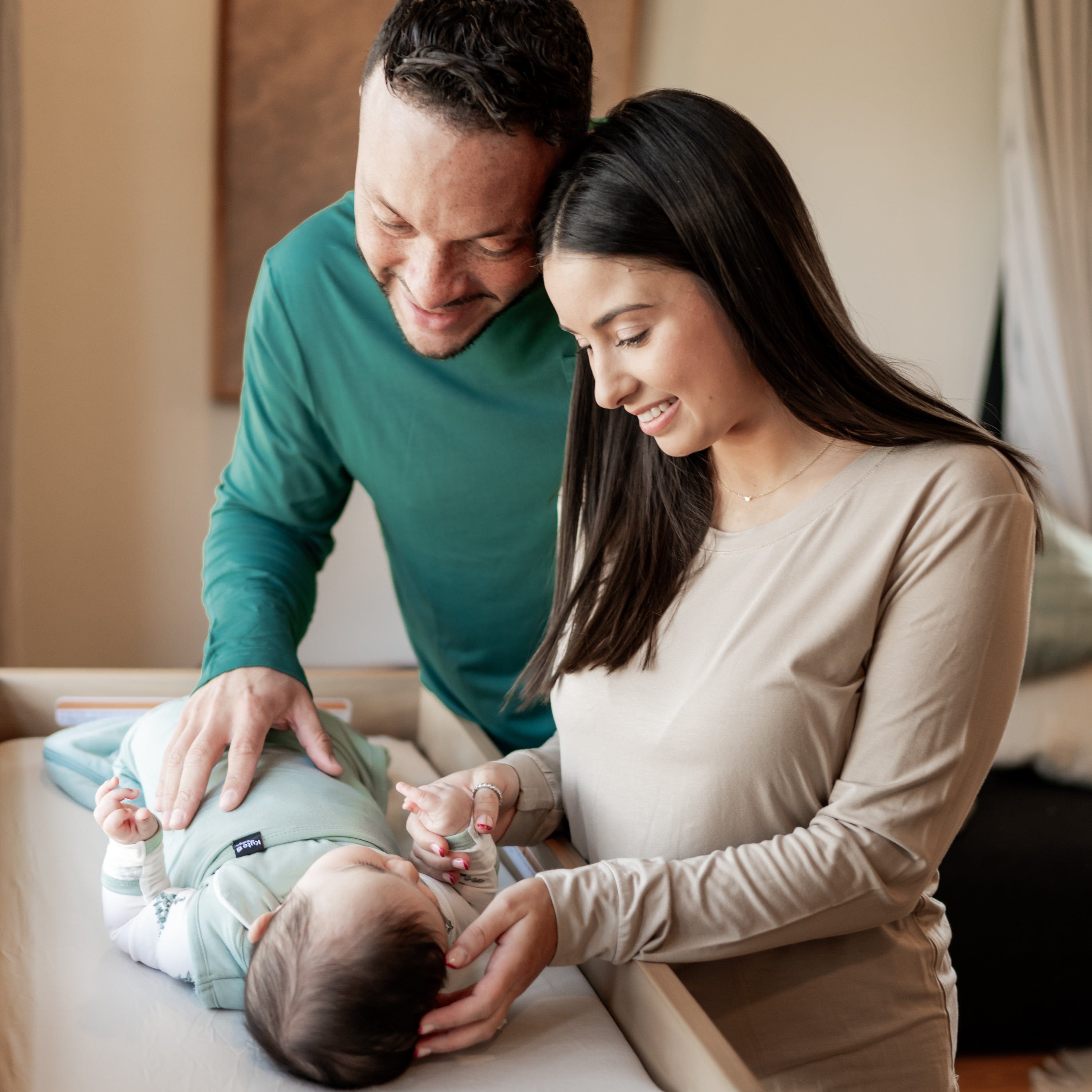 Parents standing next to bassinet as they smile down at baby wearing a Sleep Bag Swaddler in Sage