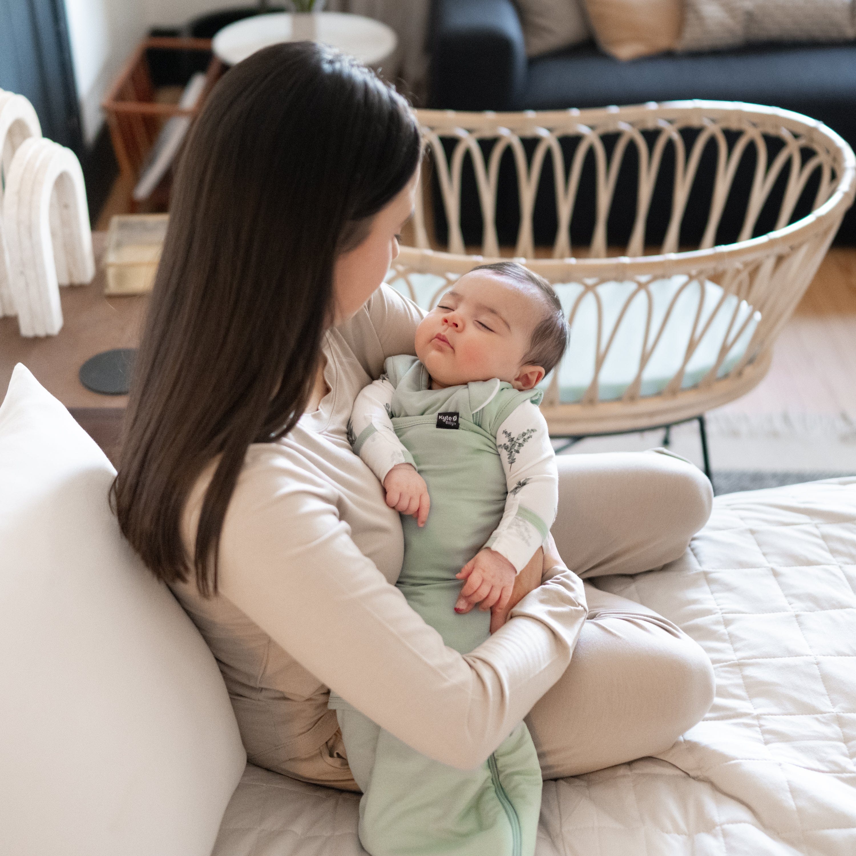 Mother sitting and holding sleeping baby while it is wearing a Sleep Bag Swaddler in Sage with bassinet in background