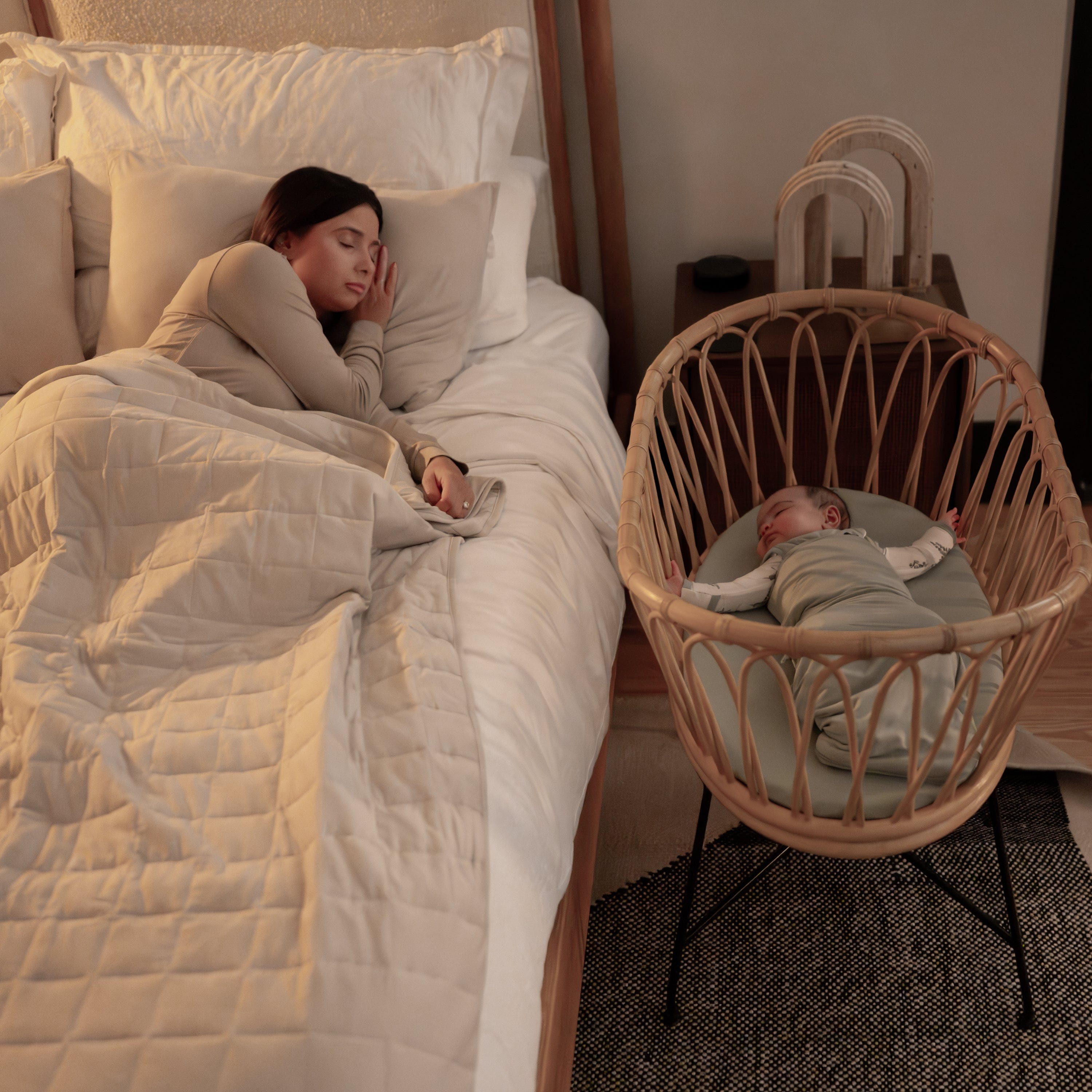 Mother sleeping in bed while baby is sleeping in rattan bassinet next to bed while wearing a Sleep Bag Swaddler in Sage