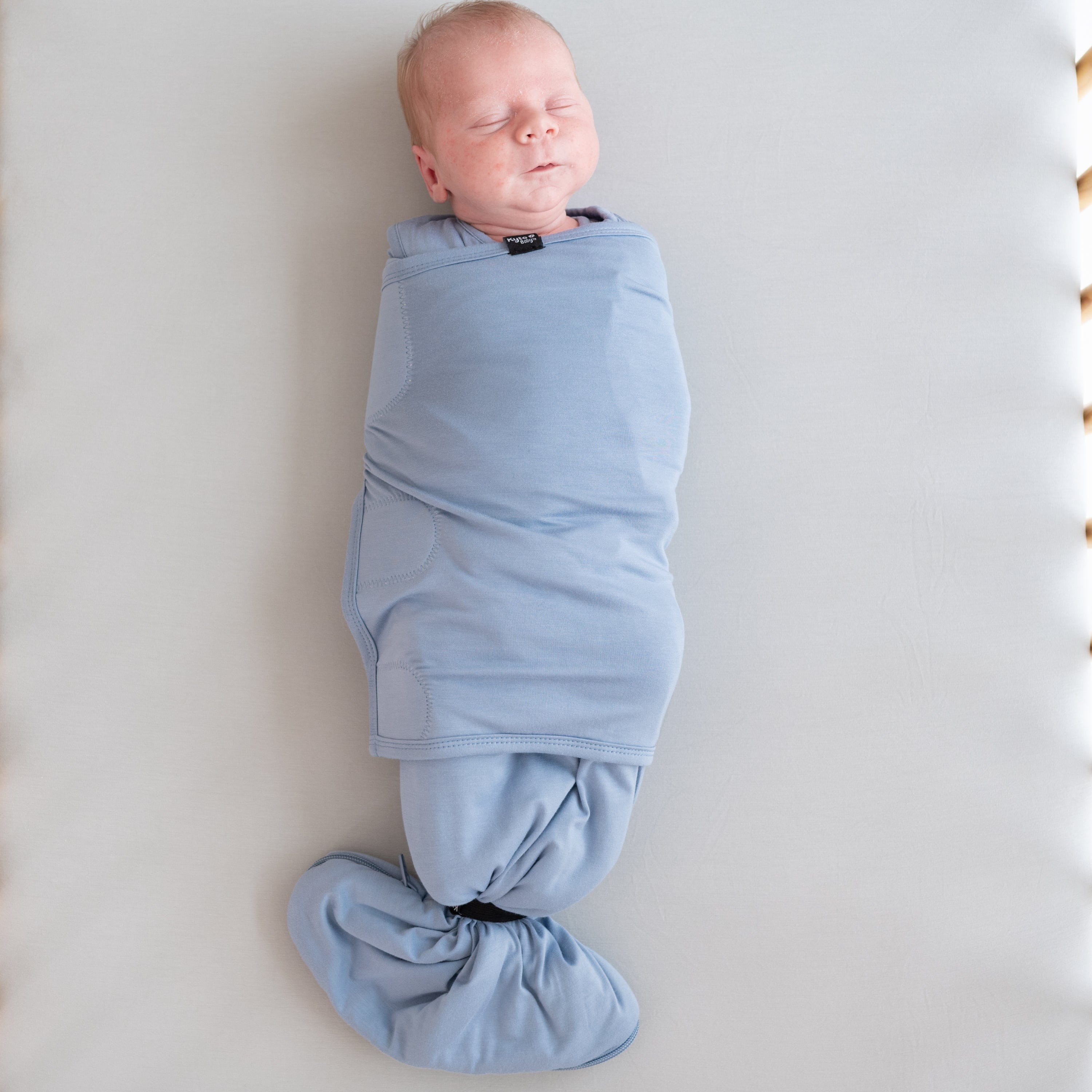 Sleeping baby wearing Kyte Baby Sleep Bag Swaddler in Slate with elastic band attached to bottom of sleep bag portion