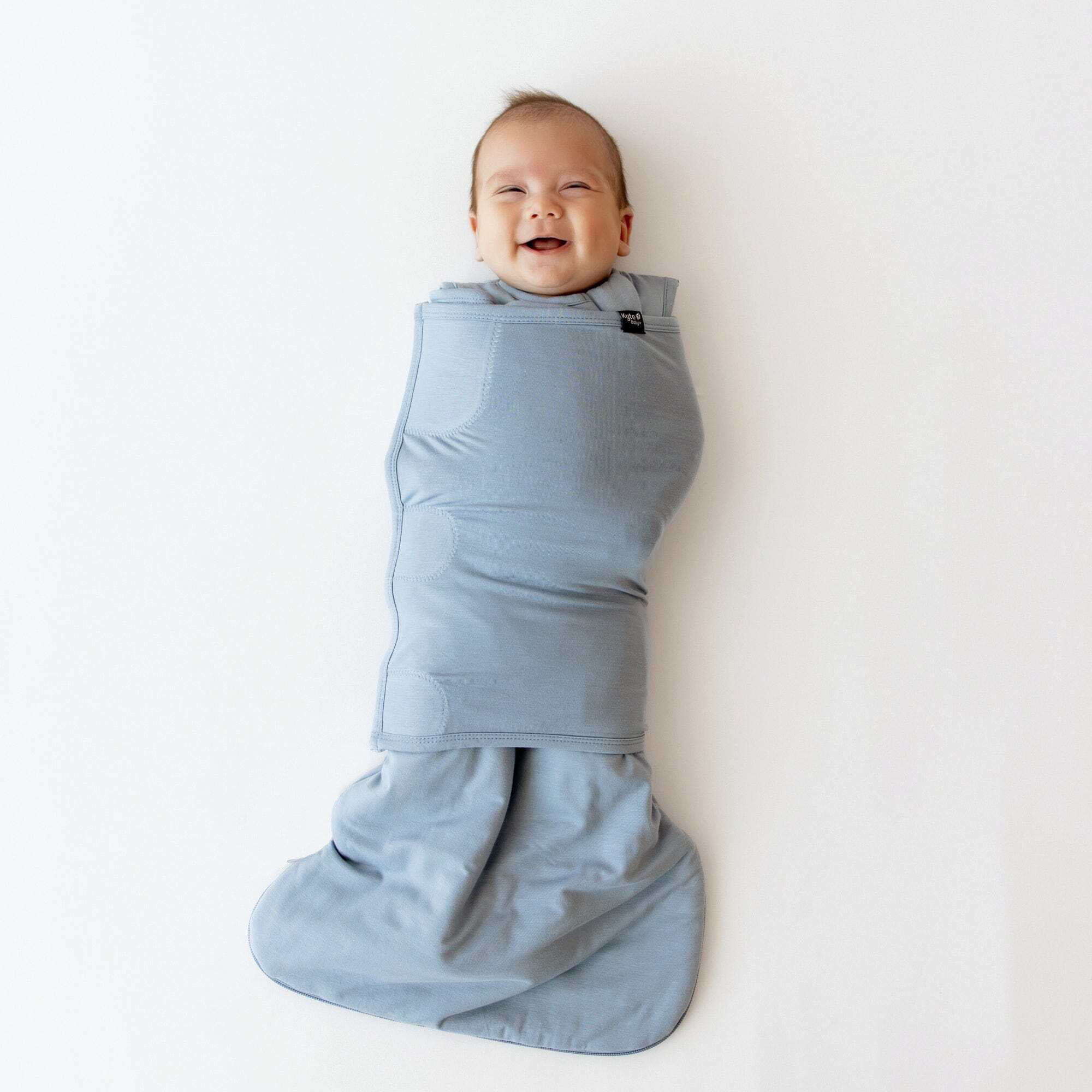 Smiling newborn laying on a blanket swaddled in the Sleep Bag Swaddler in Slate 1.0