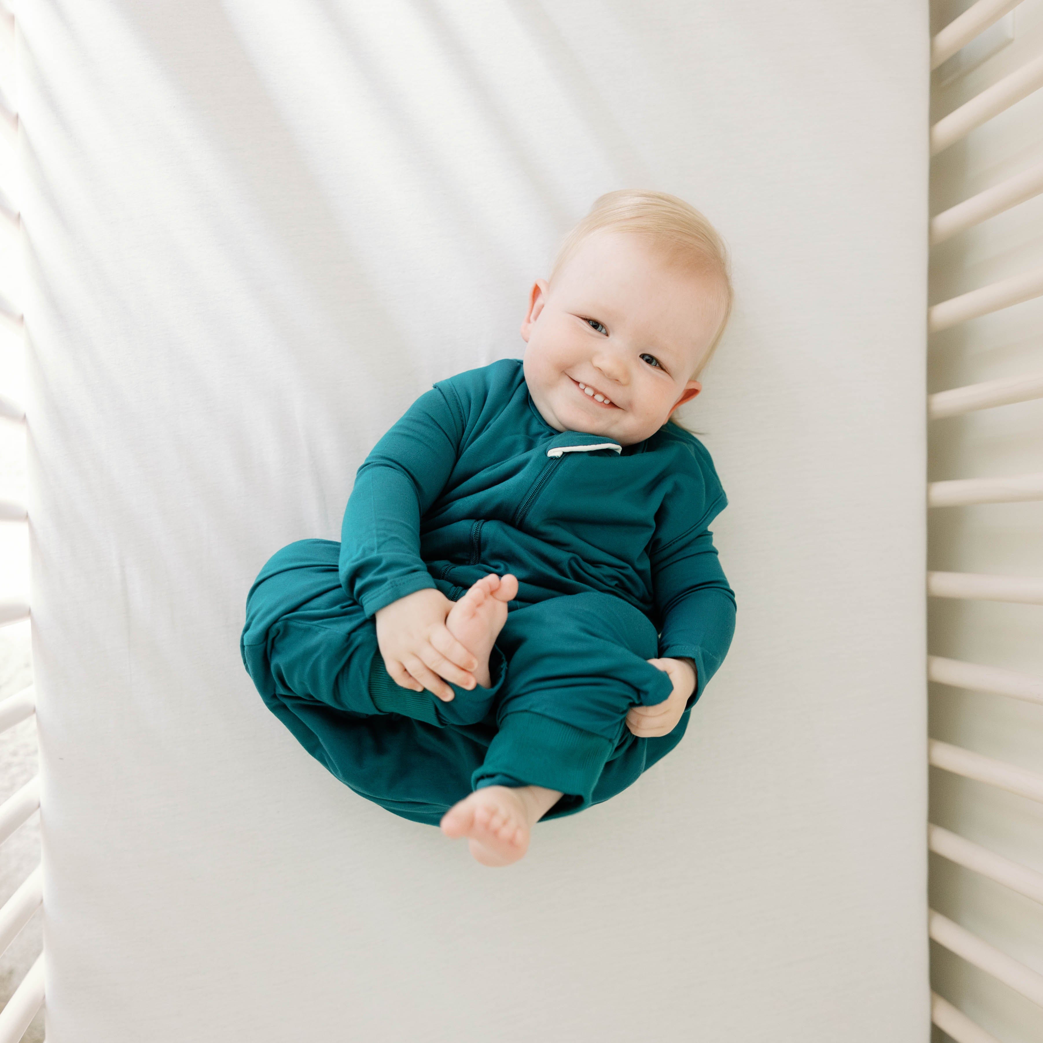 Smiling infant laying in a crib holding their leg and foot wearing the Sleep Bag Walker in Loch 1.0