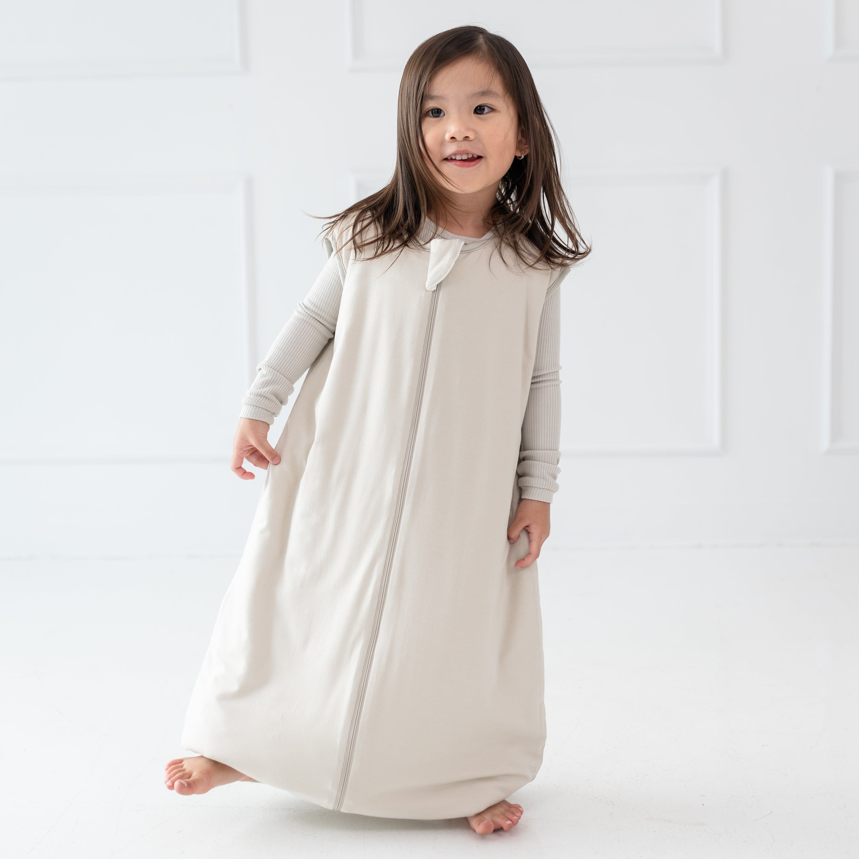 Young girl model standing on one foot wearing the Sleep Bag Walker in Oat 1.0 with matching long sleeve pajamas